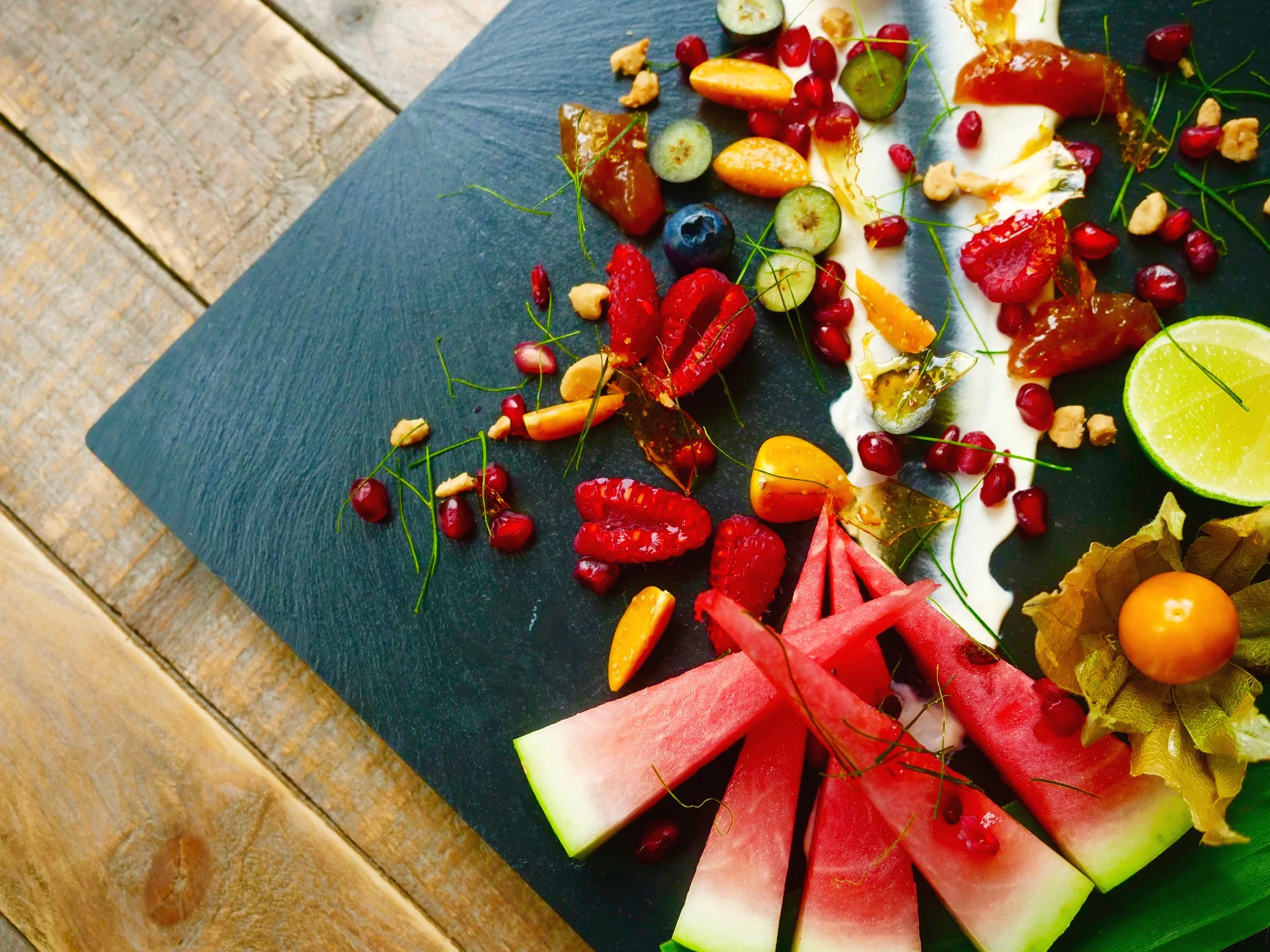 Fresh tropical fruits, pomegranate and physalis scattered over banana jam, salted peanut praline and coconut cream on a dark slate board, recipe by Sebby Holmes
