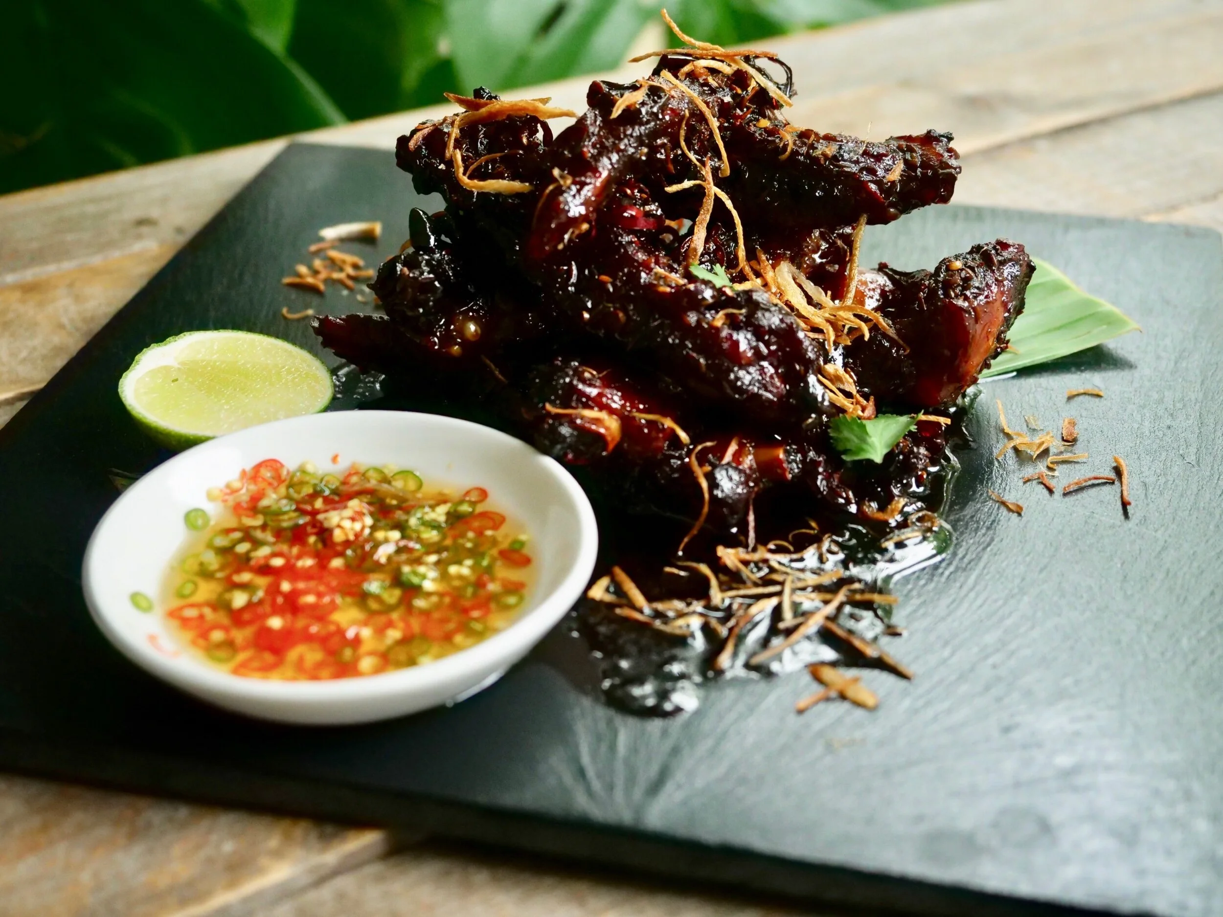 Sticky pork ribs with chilli, ginger and white pepper glaze, crispy shallots on top, prik nahm pla dipping sauce alongside on a dark slate board, recipe by Sebby Holmes