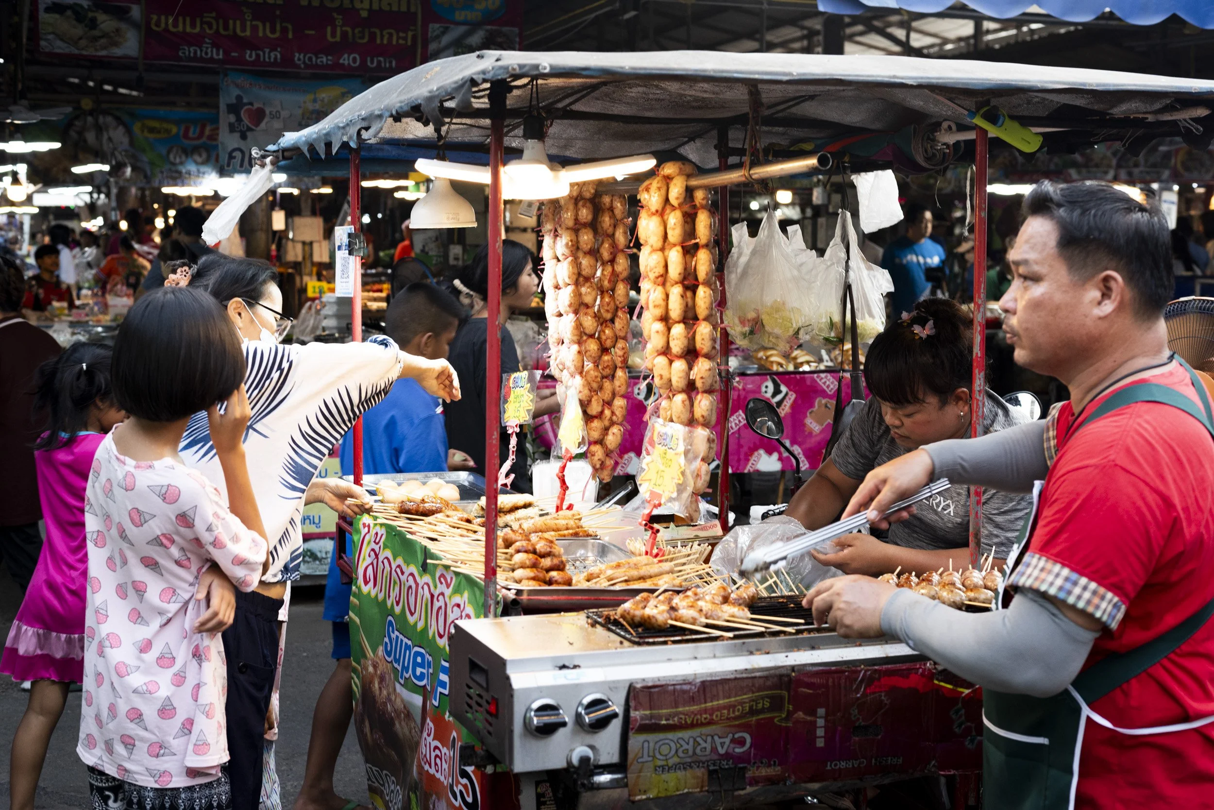 A busy Bangkok street food market at night with a vendor serving skewered snacks, photographed on a Farang trip to Thailand, by Sebby Holmes.