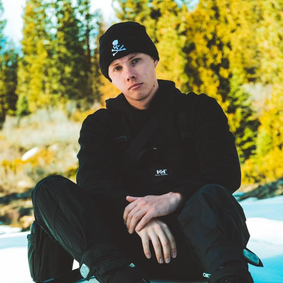Hxphy sitting outdoors in winter clothing with a black beanie, black jacket, and black pants, with a background of trees with yellow and green leaves.