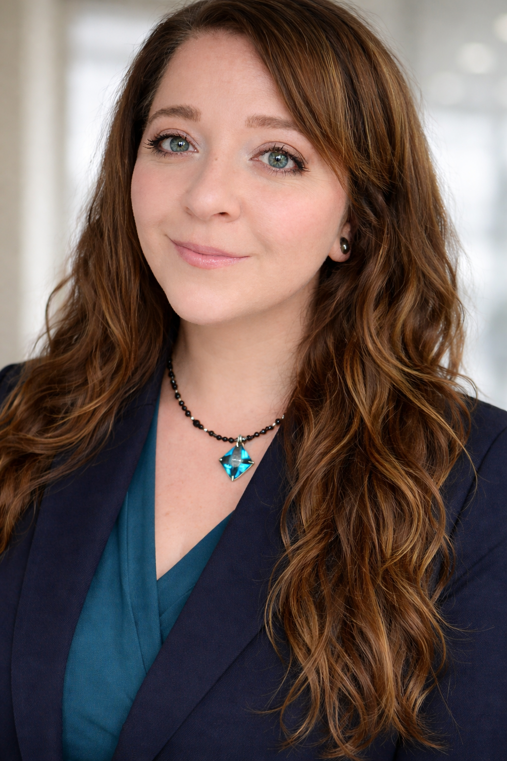 A woman with long light brown hair smiling at the camera outdoors with a blurred background of trees and water. She is wearing a black shirt with the text "You Are More Than Enough" and a necklace with a blue pendant.