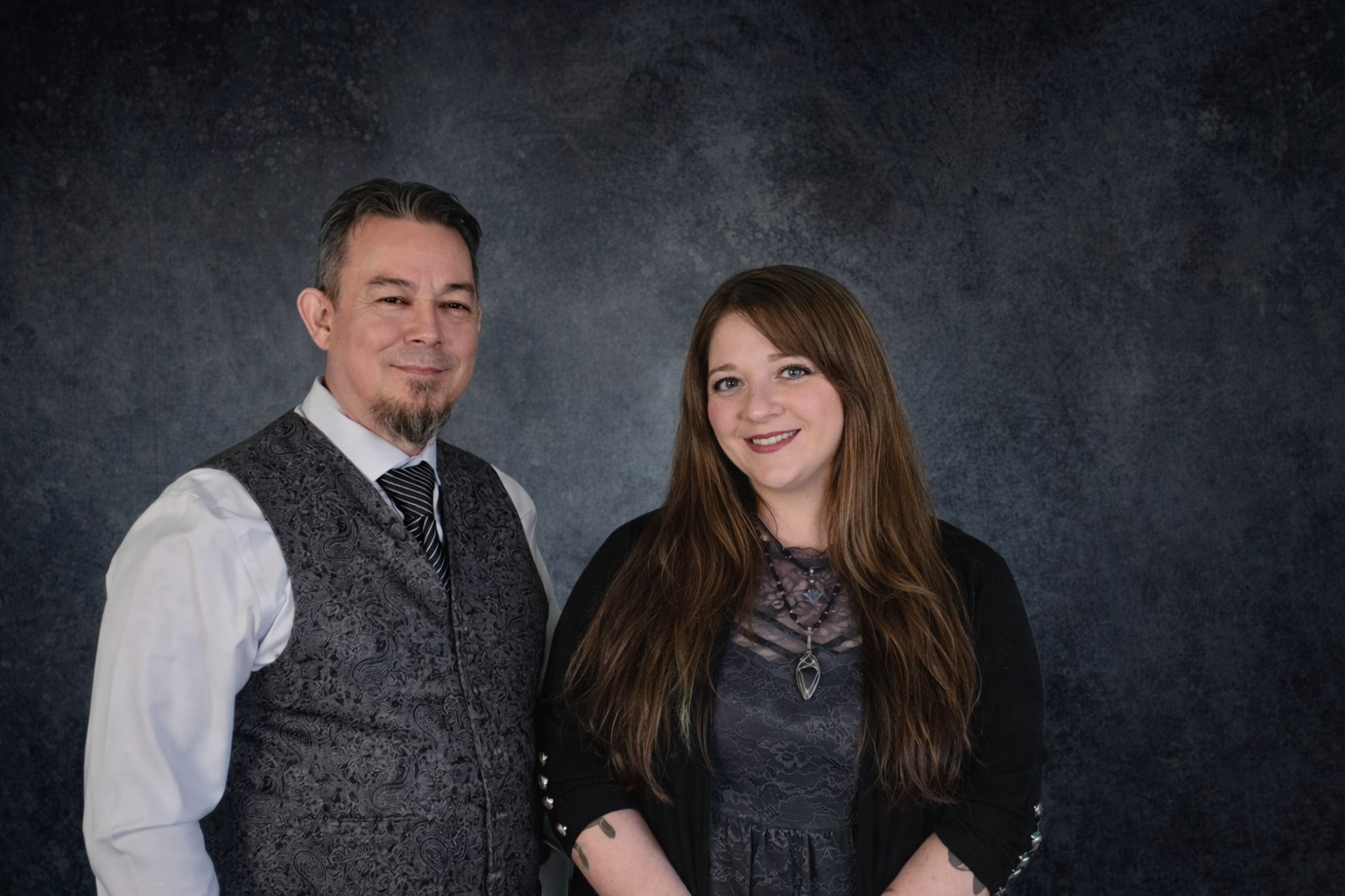 A man and a woman standing side by side, both smiling at the camera, against a dark textured background.