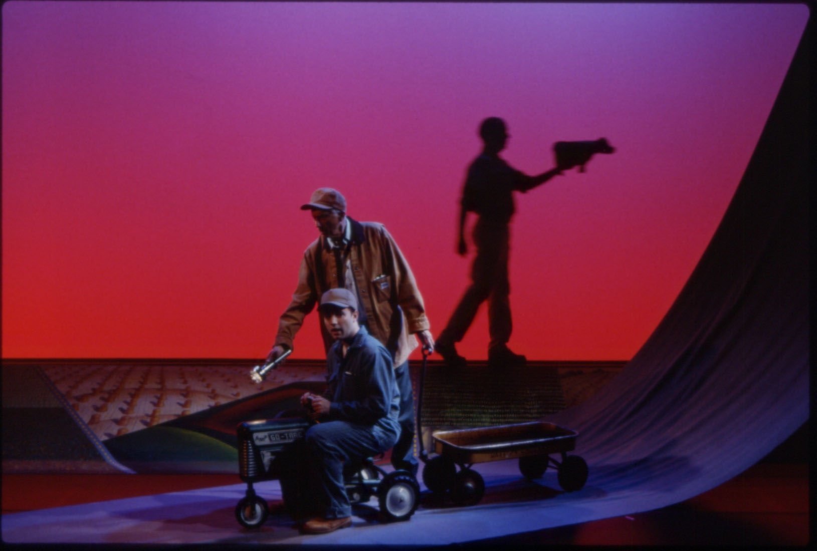 THE SHOULDER (1998) In 1994, a 73-year-old Iowa man failed his eye exam and was unable to renew his drivers license. But that didn’t discourage him from visiting his older brother in Wisconsin, who had suffered a stroke. Afraid of flying and unwilling to be driven, he embarked on an epic journey across the state of Iowa. On July 5, 1994 he packed a ten foot long trailer with camping supplies, extra clothes and gasoline, and hitched it to the back of his second-hand 1966 John Deere riding lawn mower. Achieving a top speed of 5mph, he proceeded to drive all 250 miles across the top of the state of Iowa on his lawn mower, and arrived at his brother’s farm fifty-one days later. The Shoulder is a chamber opera with music by Dan Moses Schreier, and visual design inspired by regionalist painter Grant Wood. The role of the farmer is sung simultaneously by a 60 year-old baritone and a 20 year old tenor highlighting the issues of aging, youthful spirit, and the passage of time that are inherent to the story. Hurlin plays the state of Iowa. Performed by Don Chastain, Dan Hurlin, Doug Marcks Music composed by Dan Moses Schreier Music direction and performance by Alan Johnson Set design by Dan Hurlin Lights by Tyler Micoleau Premiered at Dance Theater Workshop, NYC Toured nationally AWARD : American Theater Wing Design Nomination Photos: Tyler Micoleau