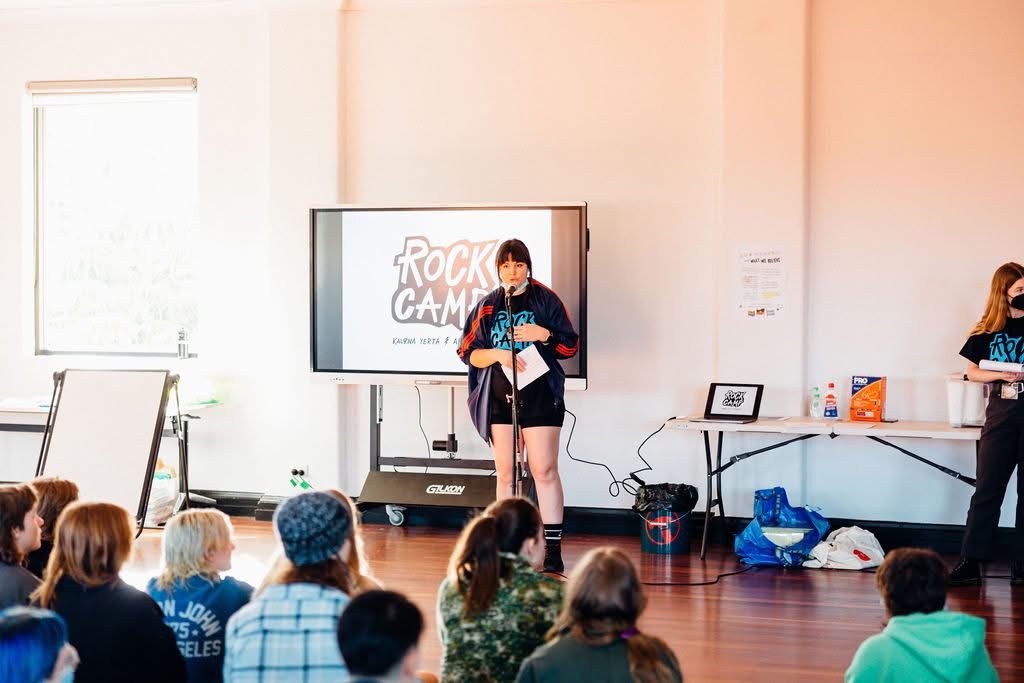 A girl standing on stage speaking into a microphone during a presentation at a camp, with an audience of children seated on the floor watching her.