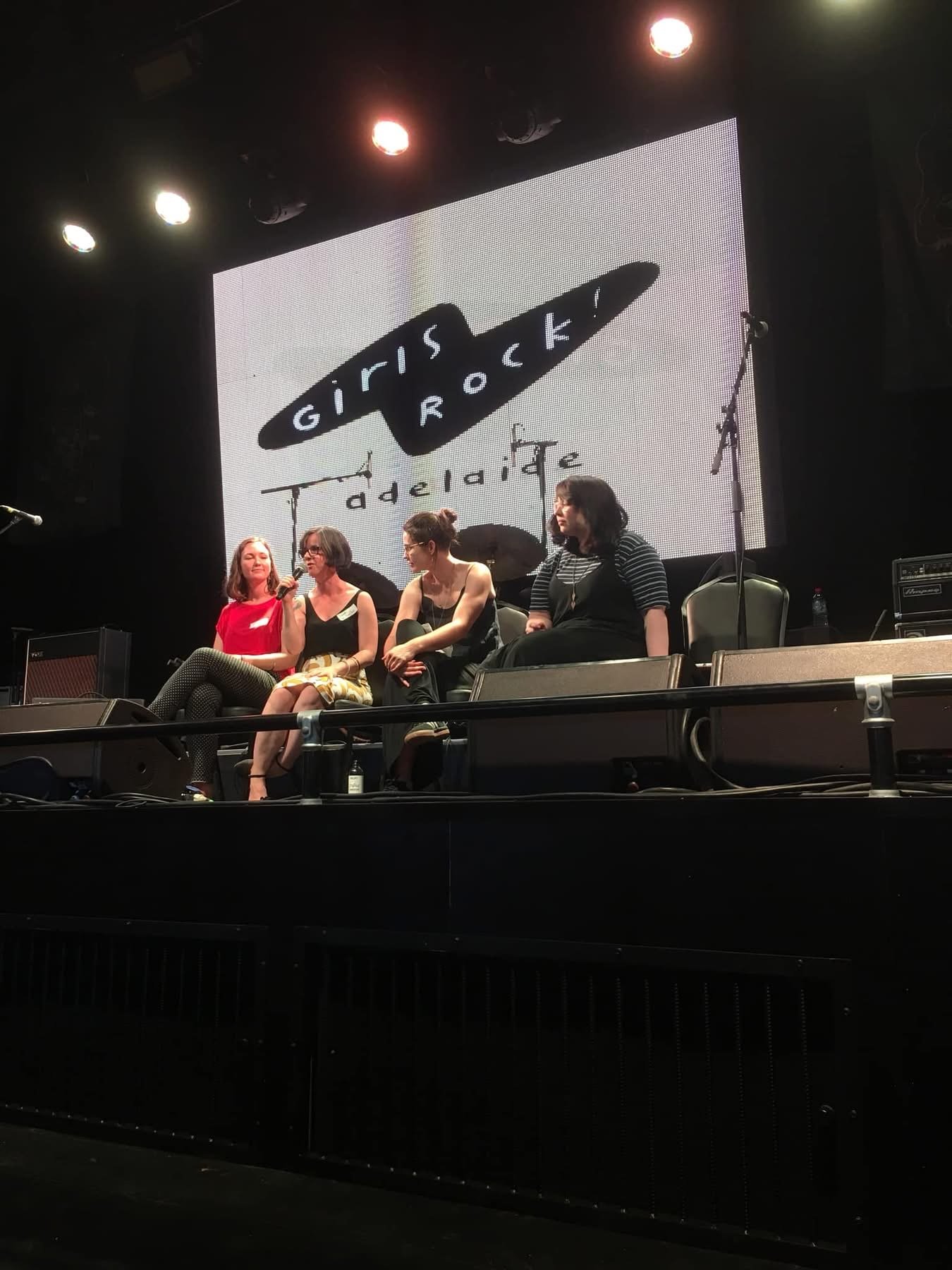 Four women are sitting on stage in front of a large screen that displays the logo for 'Girls Rock! Adelaide', including Anna jeavons. One woman, Hannah Fairlamb is holding a microphone and talking, while the others listen.