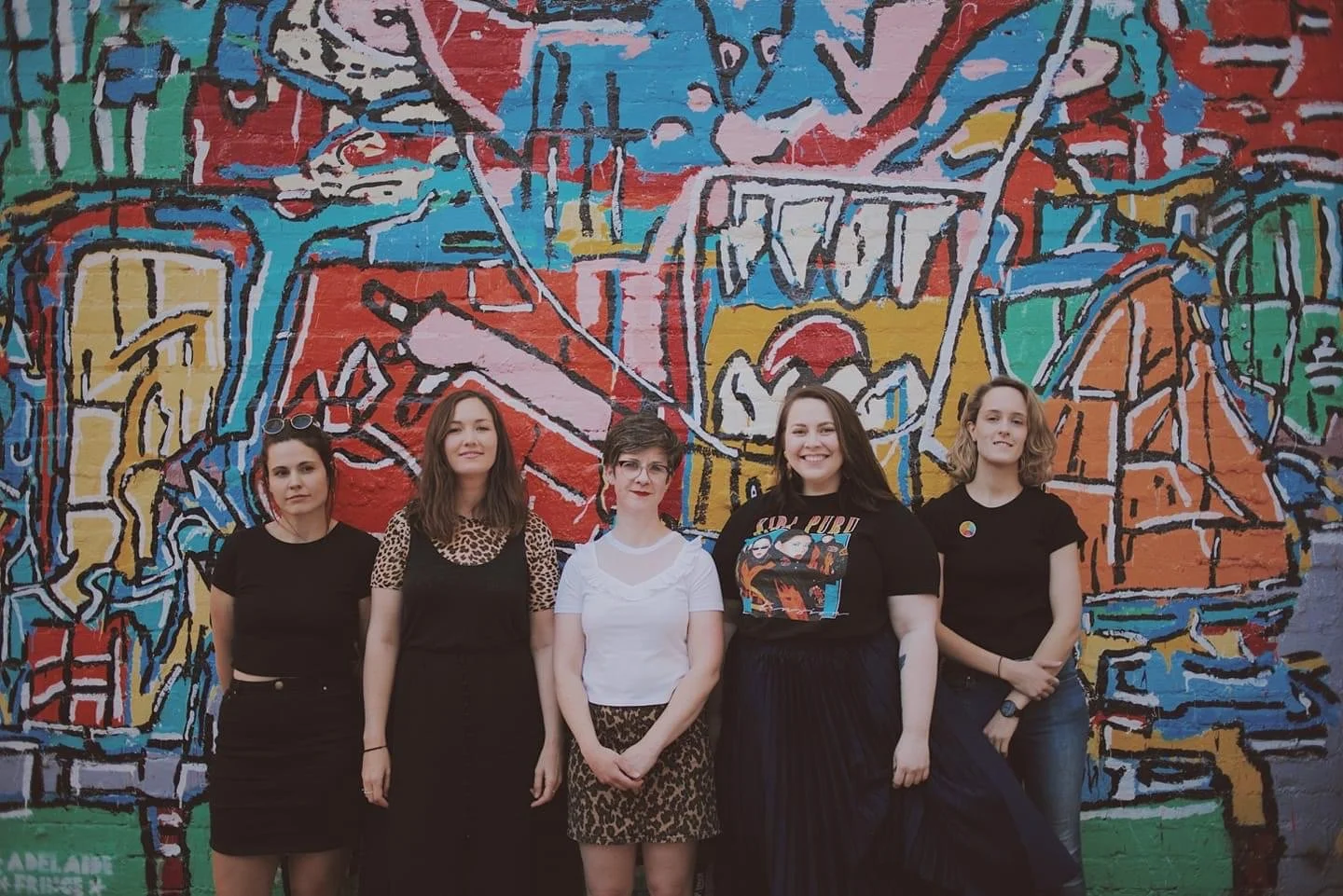 Five people are standing in front of a colourful graffiti mural on a brick wall. They are the Girls Rock! Adelaide committee. 