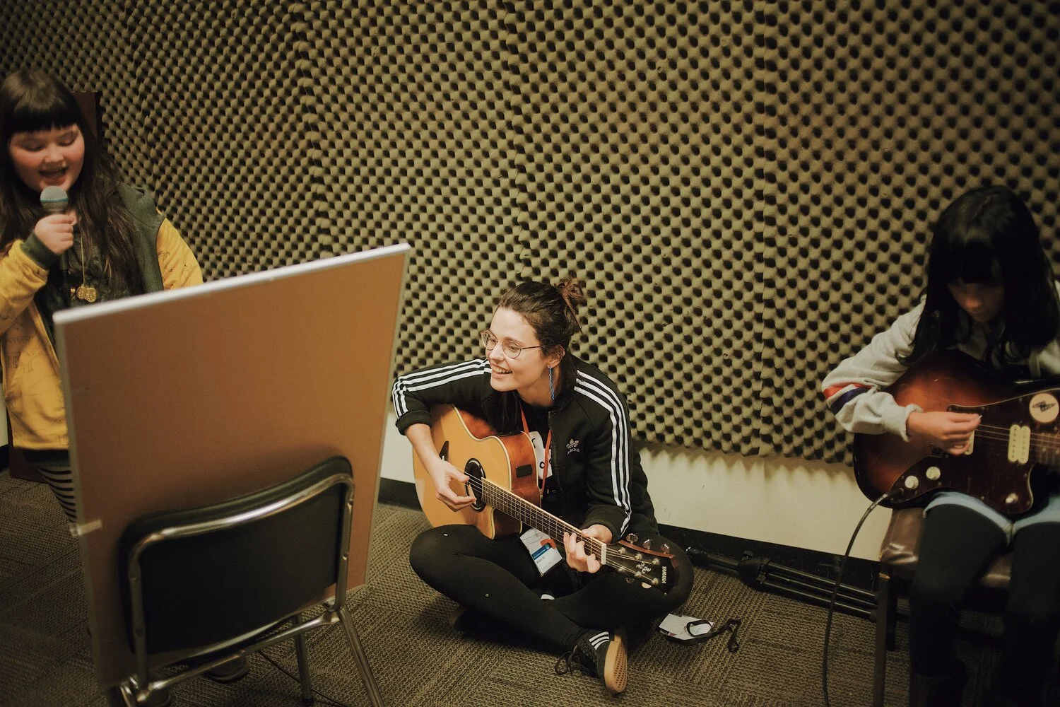 Anna Jeavons sitting on the floor playing an acoustic guitar and smiling, with a young girl singing into a microphone on the left and another girl playing an electric guitar on the right, in a room with soundproofing foam on the wall.