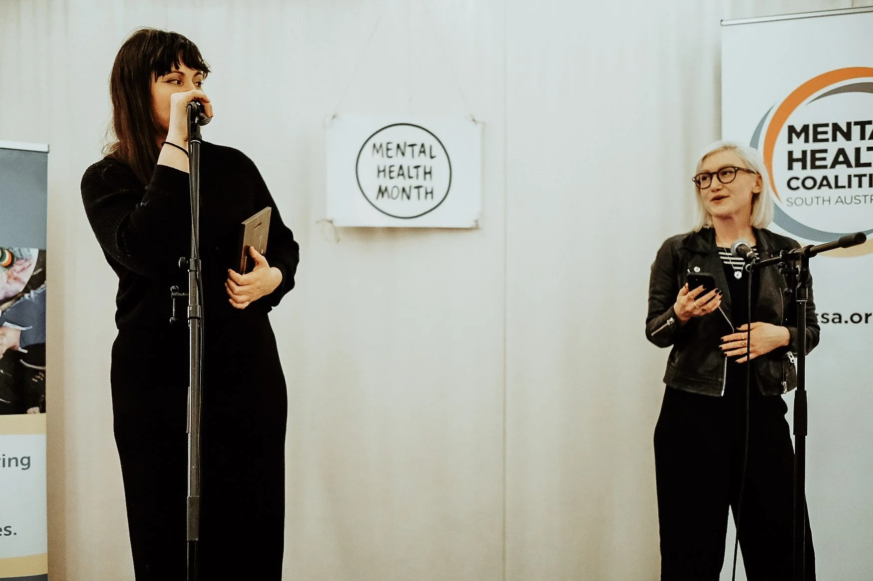 Two women speaking at a mental health awareness event, with signs reading "Mental Health Month" and "Mental Health Coalition South Australia" in the background. Anna Jeavons speaks into the microphone. 