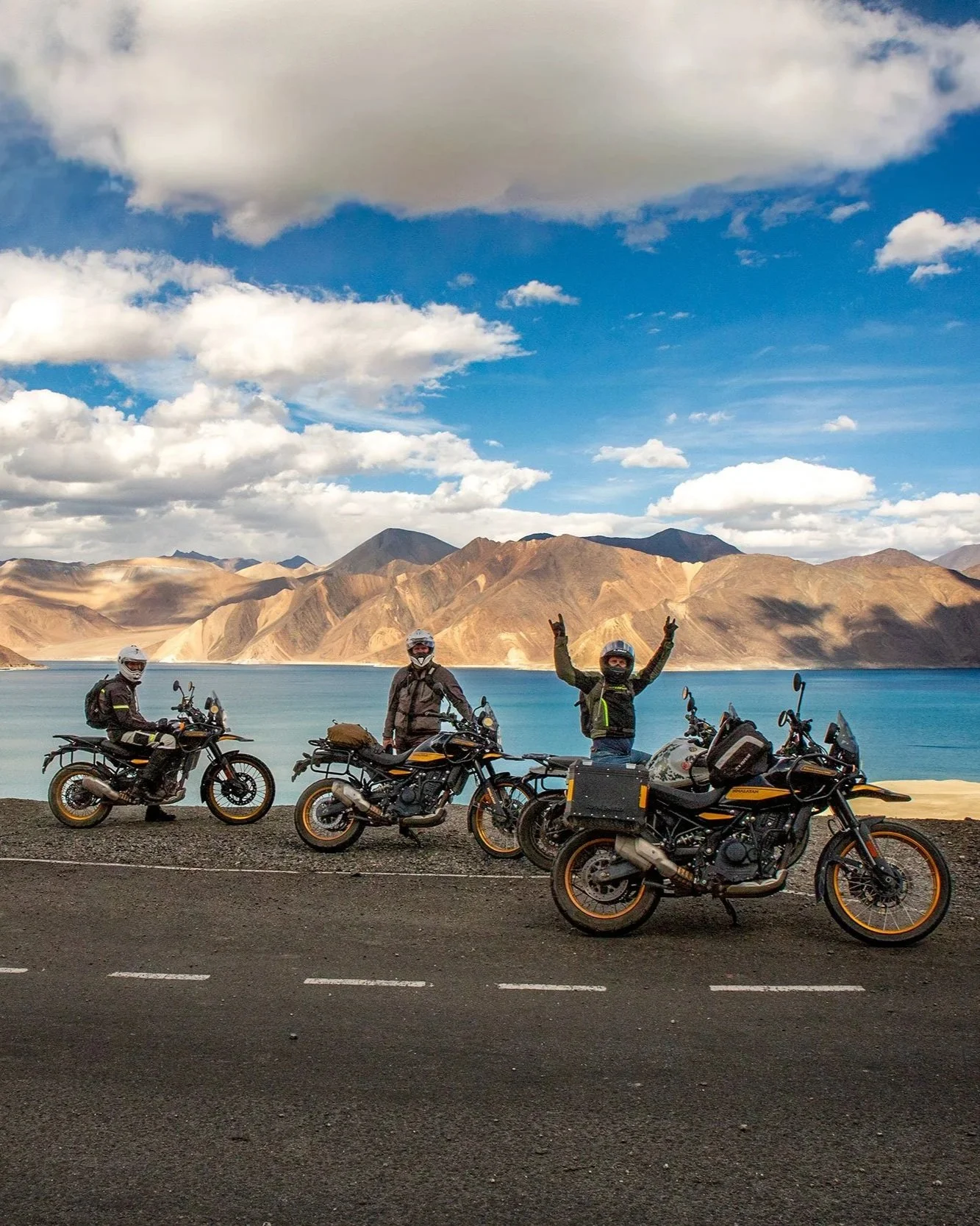 Three men posing with bikes on a road near a lake and mountains under a partly cloudy sky.