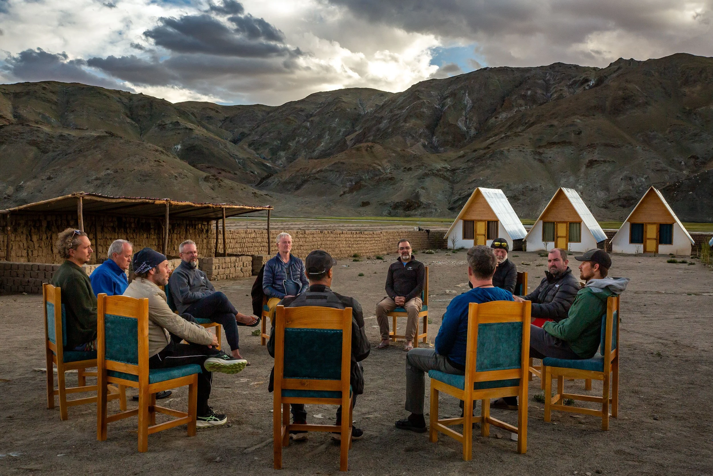 A group of men sitting in chairs outdoors in a men's circle, with a backdrop of mountains and a cloudy sky, possibly taking part in a group discussion.