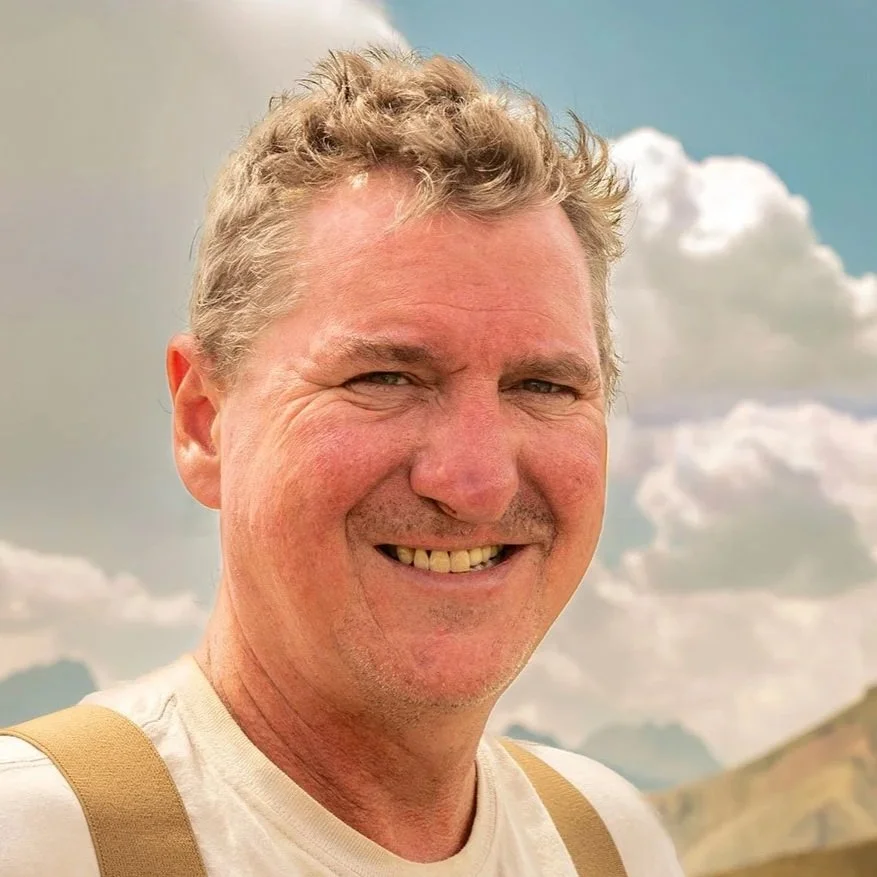 A smiling middle-aged man with short curly hair, wearing a light-colored shirt and suspenders, standing outdoors with a cloudy sky and hills in the background.