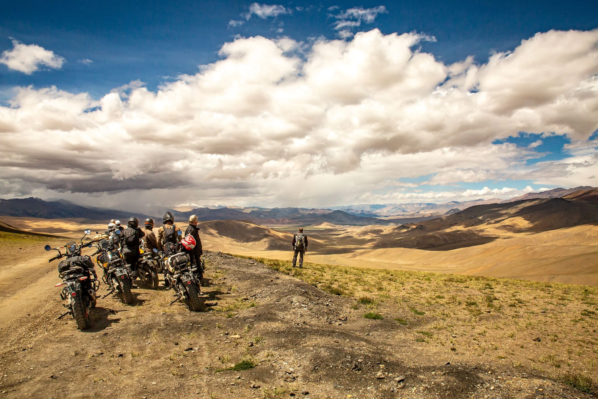 A group of men with gear and helmets are parked on a dirt path contemplate the vast mountainous landscape under a partly cloudy sky.