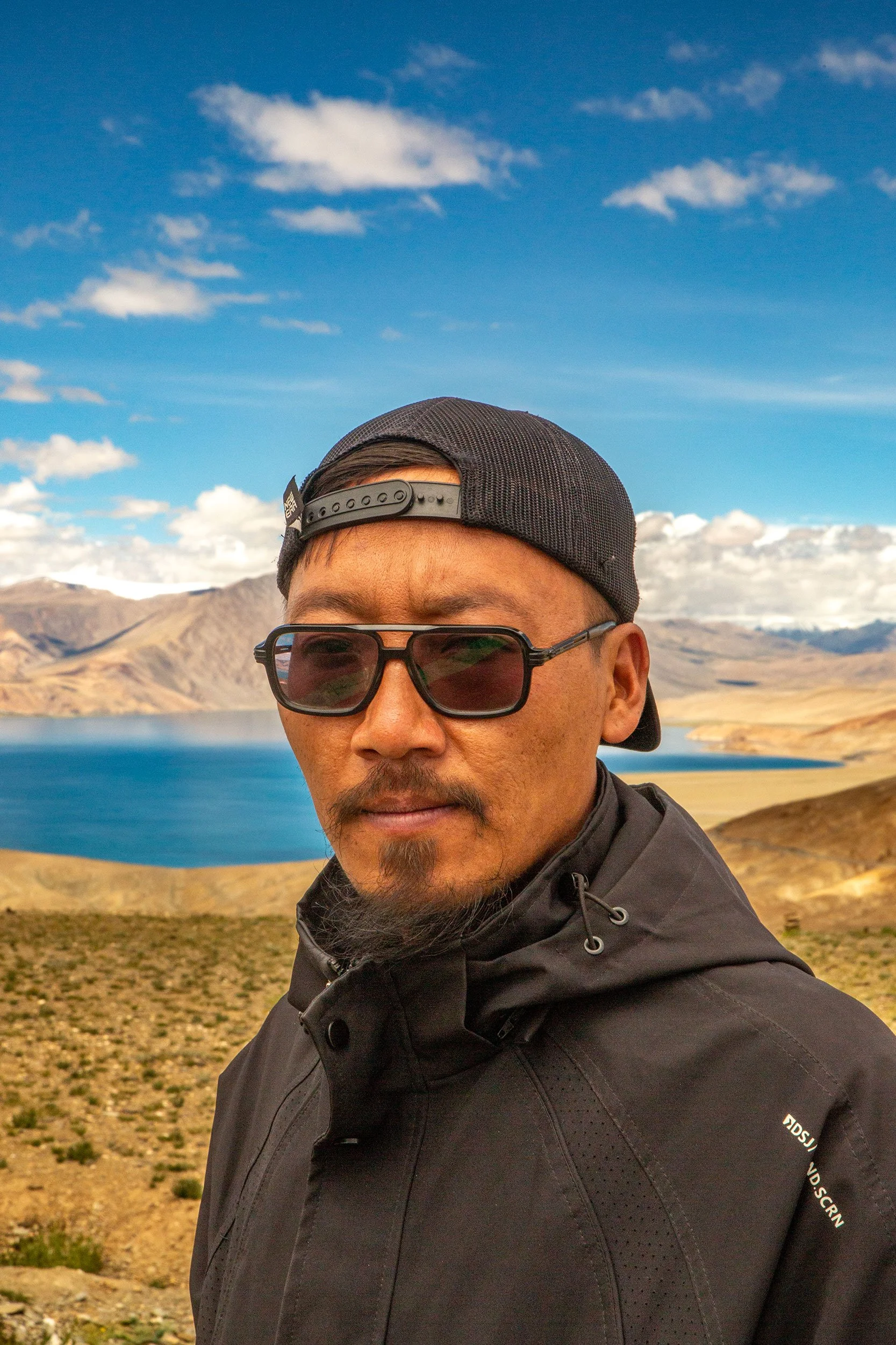 A man wearing sunglasses and a black cap with an outdoor landscape behind him, including mountains, a lake, and partly cloudy sky.