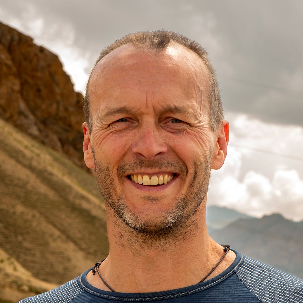 Close-up of smiling man outdoors with mountains and cloudy sky in the background.
