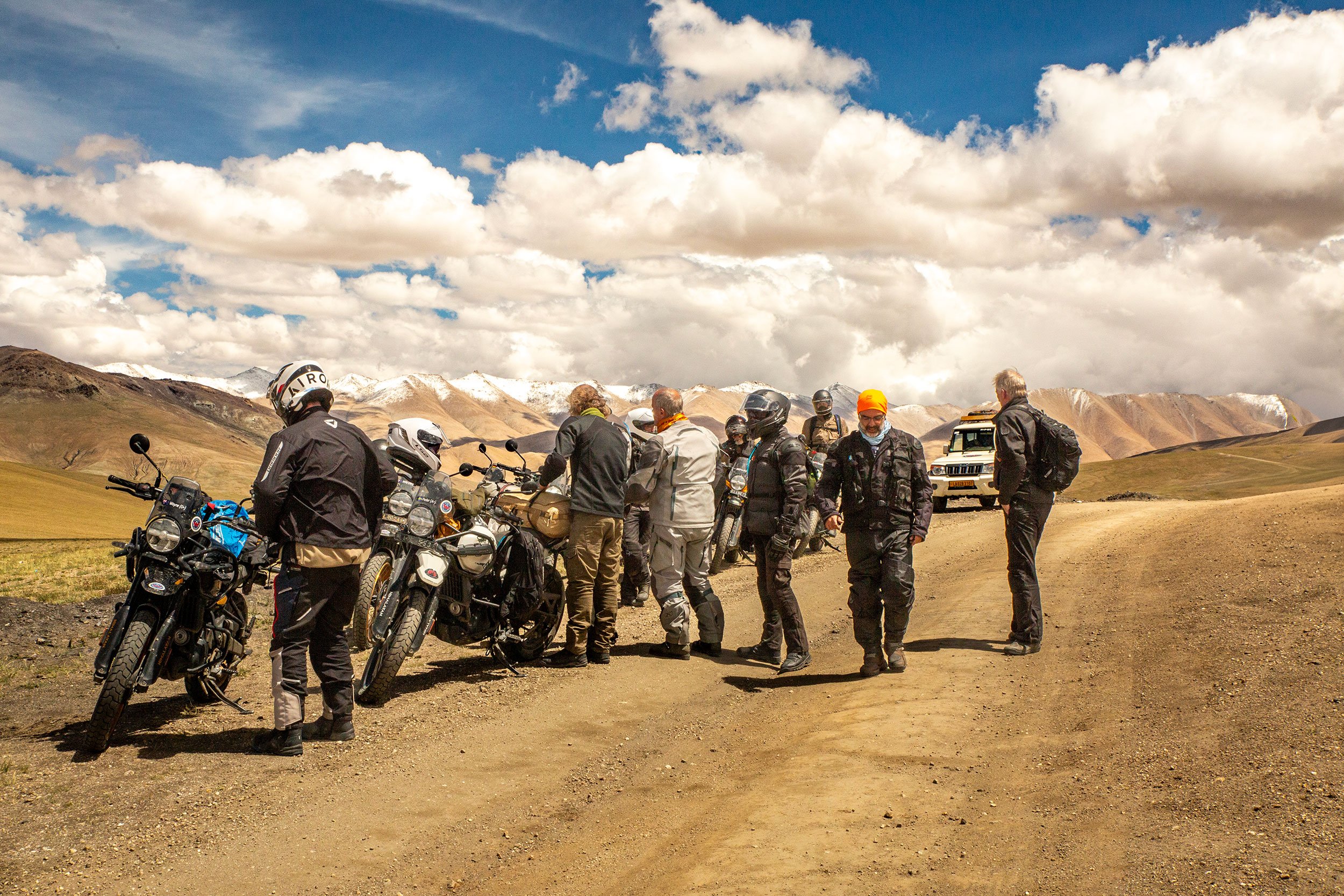Group of men gathered outdoors in a mountainous landscape with partly cloudy skies, some snow-capped peaks in the background, and a dirt path.