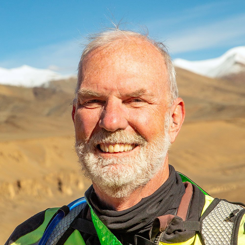 A smiling elderly man with white hair and beard, wearing motorcycle gear, standing in a mountainous landscape with snow-capped peaks in the background on a clear day.