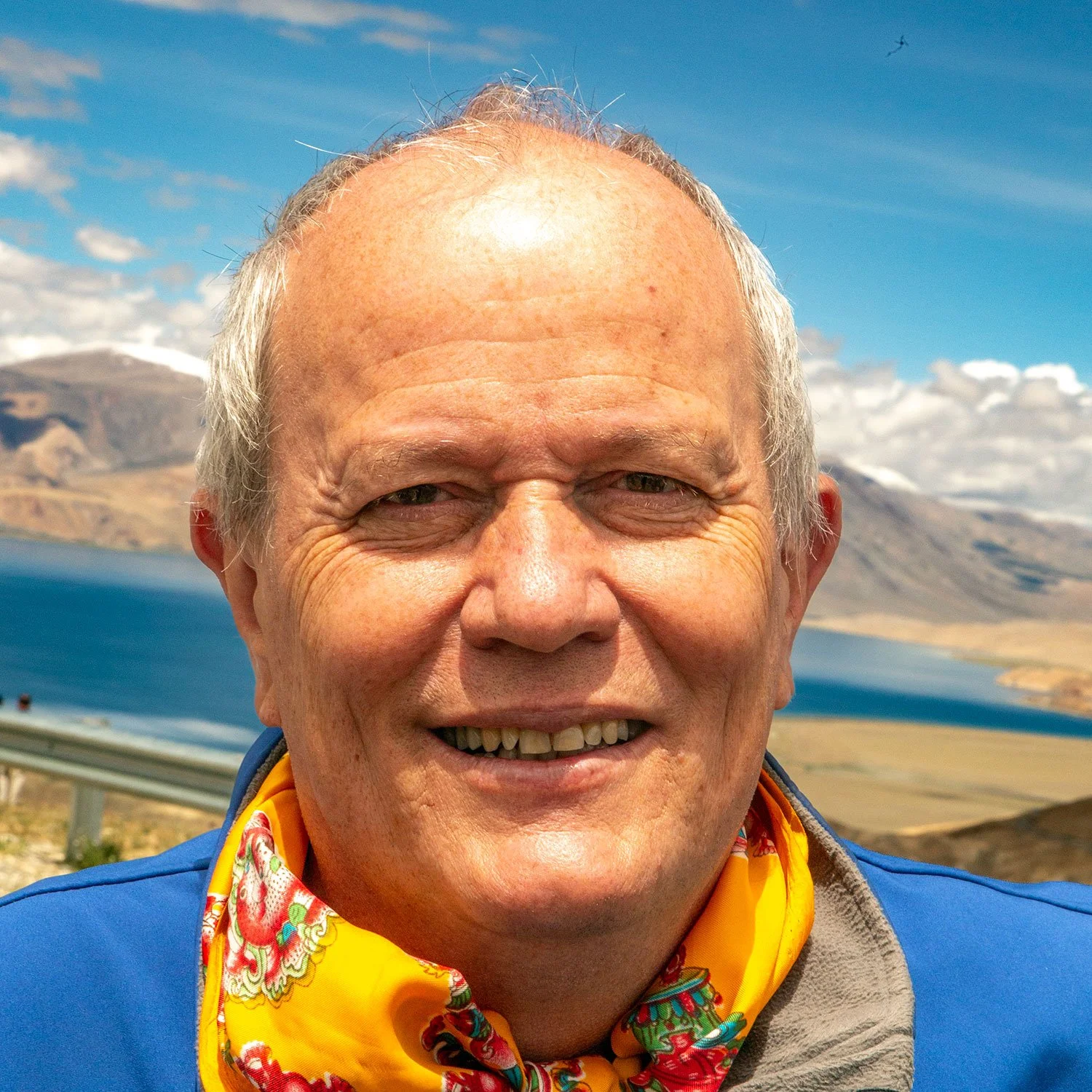 Close-up of smiling elderly man wearing a colorful neckerchief, blue jacket, outdoors with mountains, lake, sky and clouds in the background.