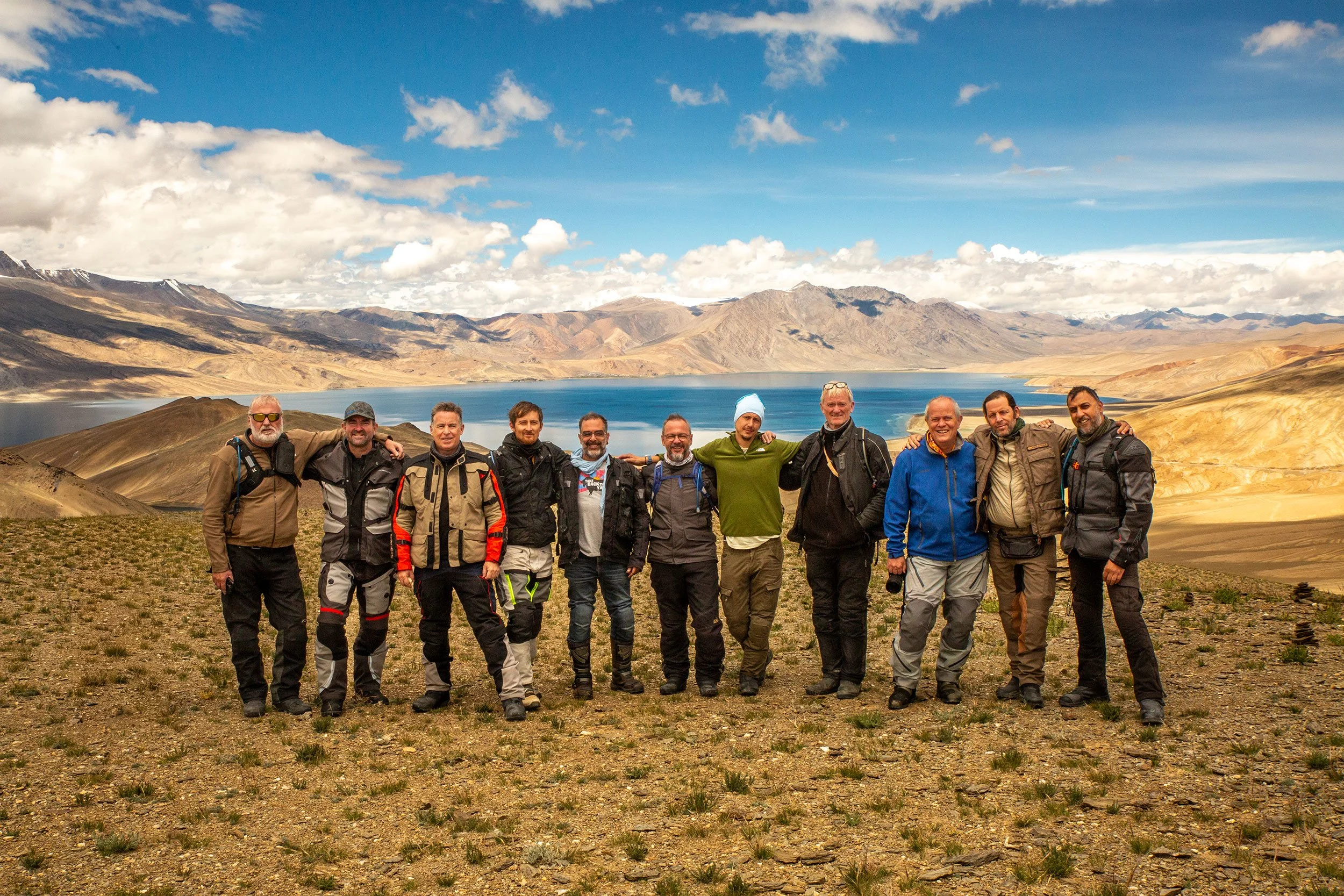 A group of eleven men standing together outdoors in a mountainous landscape with a large lake, rugged hills, and a partly cloudy sky in the background.
