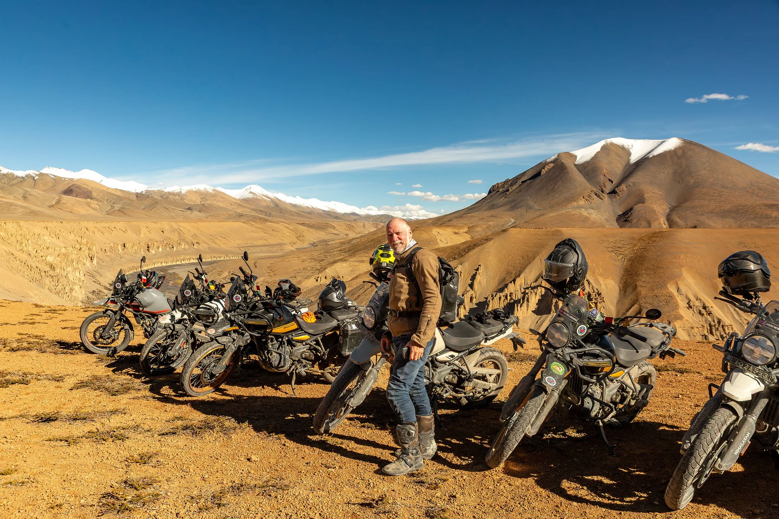 A man stands next to a line of motorcycles in a desert landscape with a mountain in the background under a blue sky.