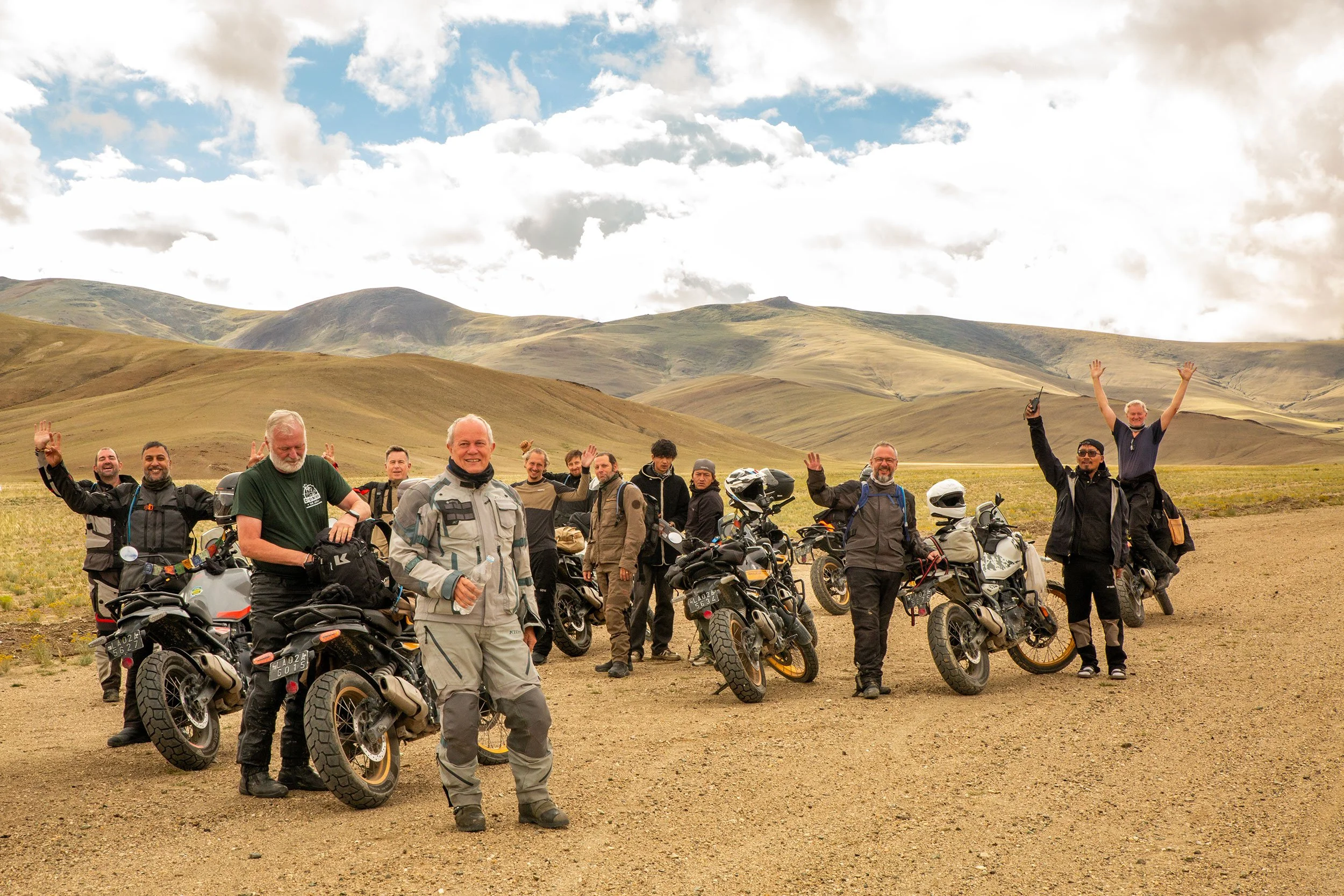 A group of men posing with their bikes in a desert landscape with rolling hills and partly cloudy sky.