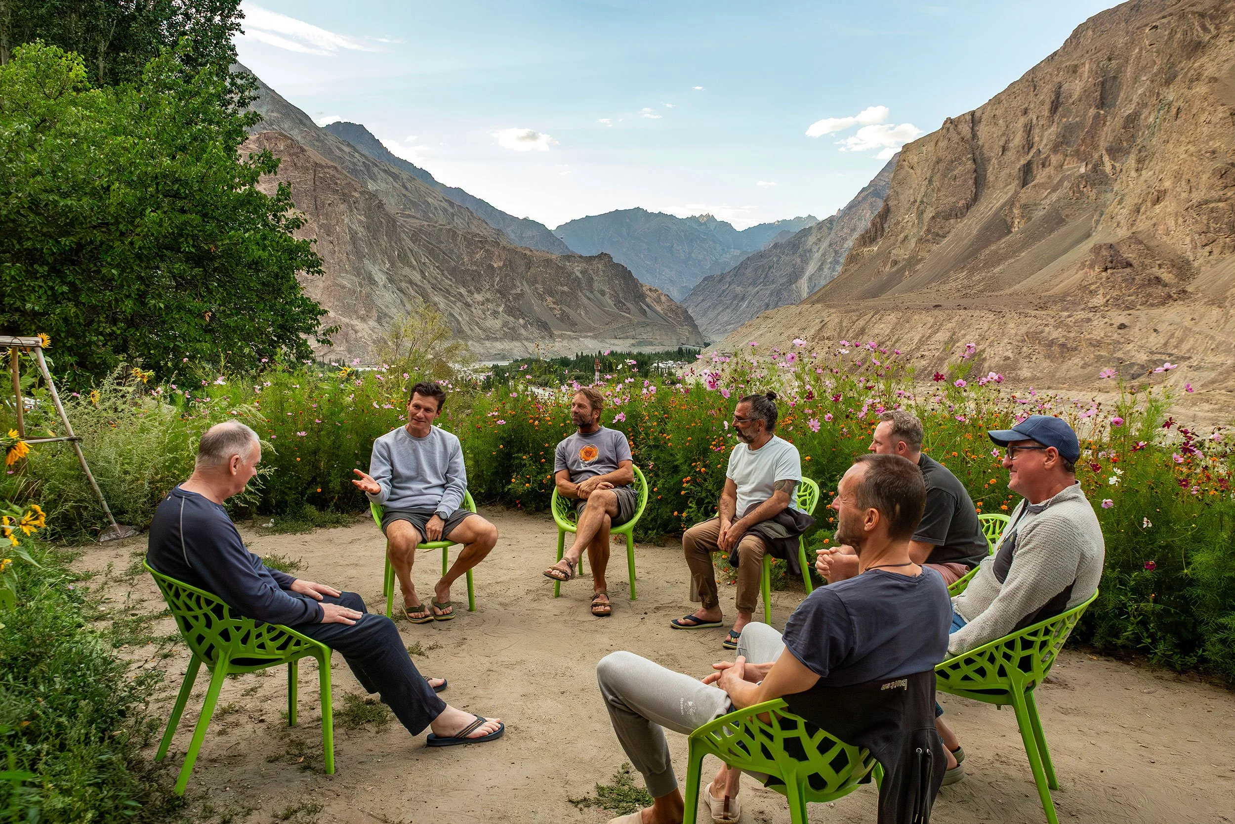 A group of men sitting in a circle outdoors in front of a mountainous landscape with colorful wildflowers in the foreground. One man gives feedback to another, as the rest listens to him.