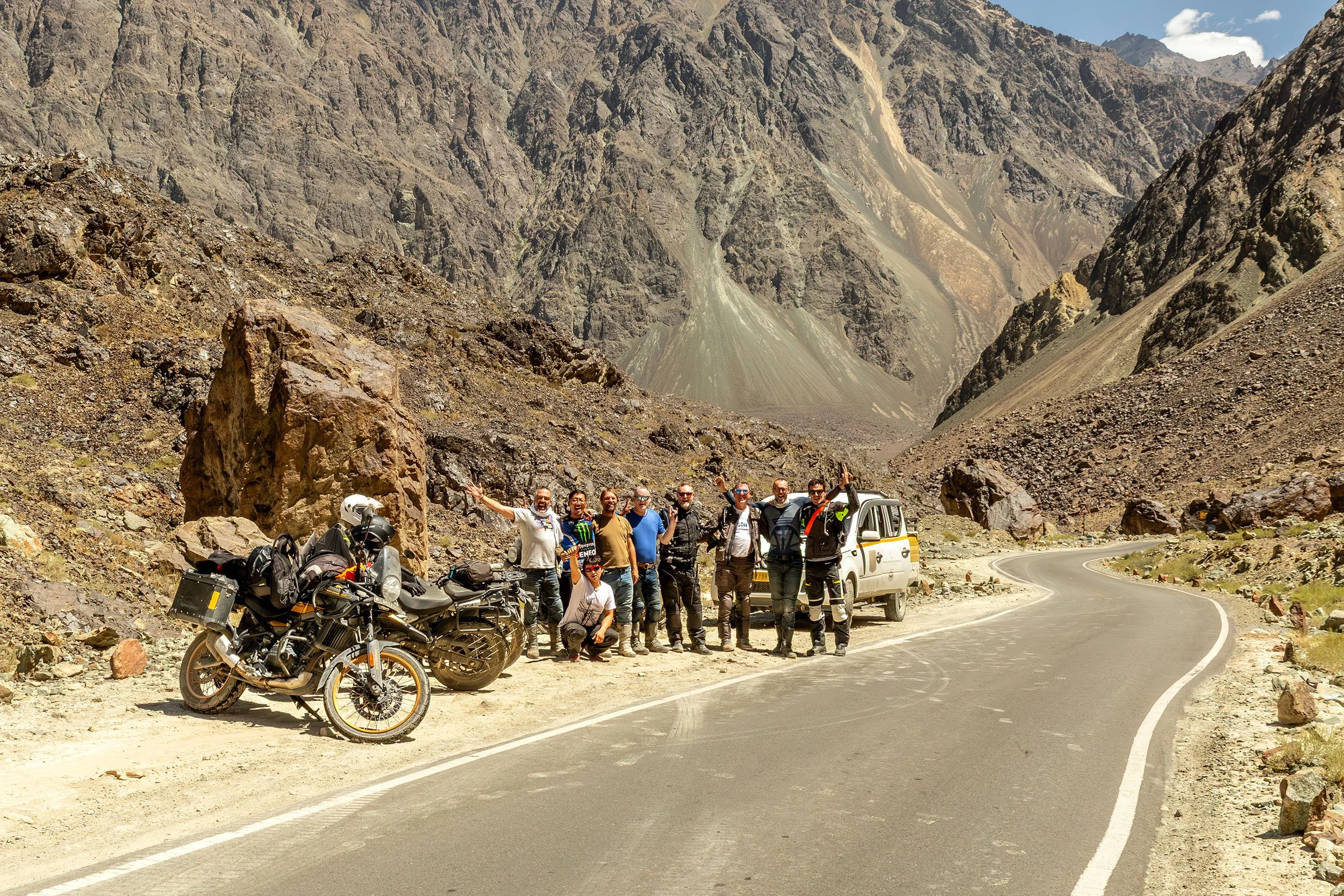 Group of people with motorcycles and a vehicle standing on the side of a winding mountain road with rocky cliffs and mountains in the background.