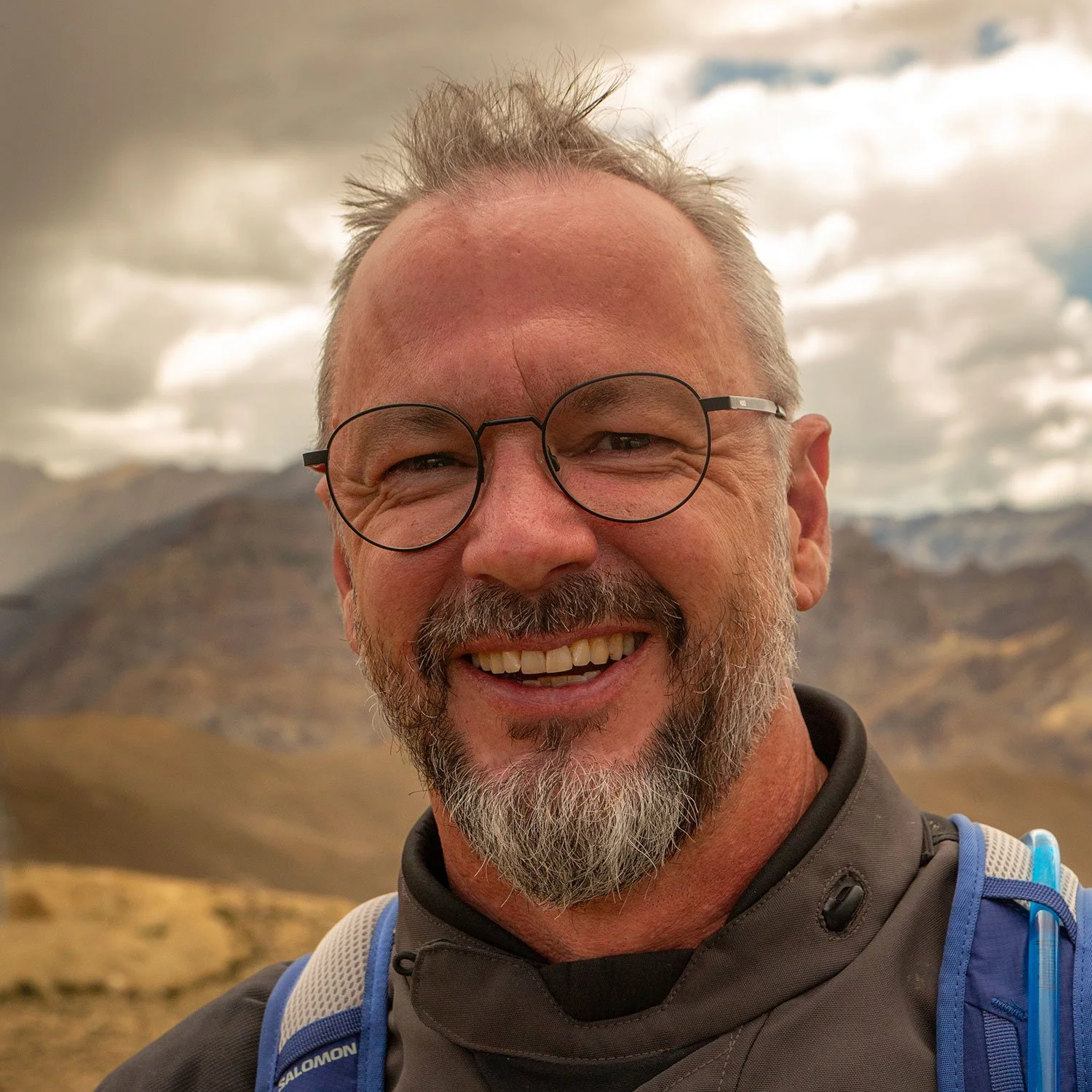 A smiling middle-aged man with glasses, a beard, and spiky hair, outdoors in a mountainous landscape under cloudy skies.