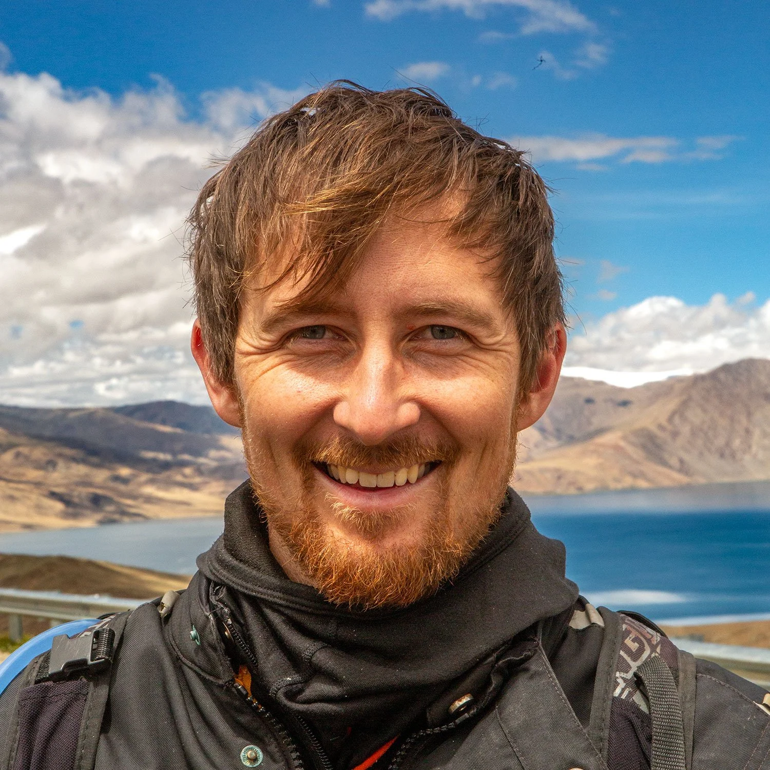 A smiling man with light brown hair and a beard outdoors, with a mountain landscape, lake, and cloudy sky in the background.