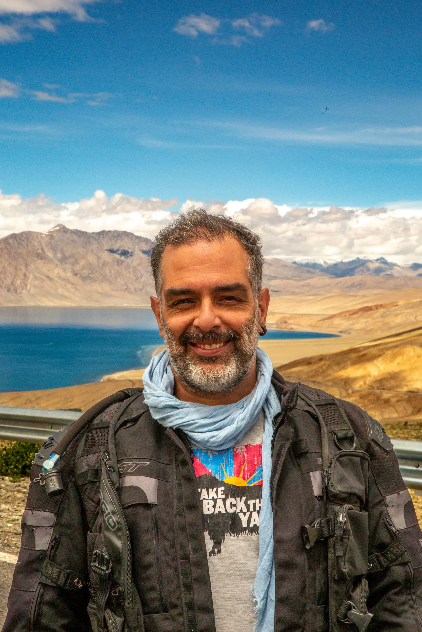 A smiling man with short hair and a beard standing outdoors near a lake with mountains in the background. He is wearing a black motorcycle jacket, a graphic t-shirt, and has a light blue scarf around his neck. A backpack is visible on his shoulders, and there is an airplane flying in the sky.