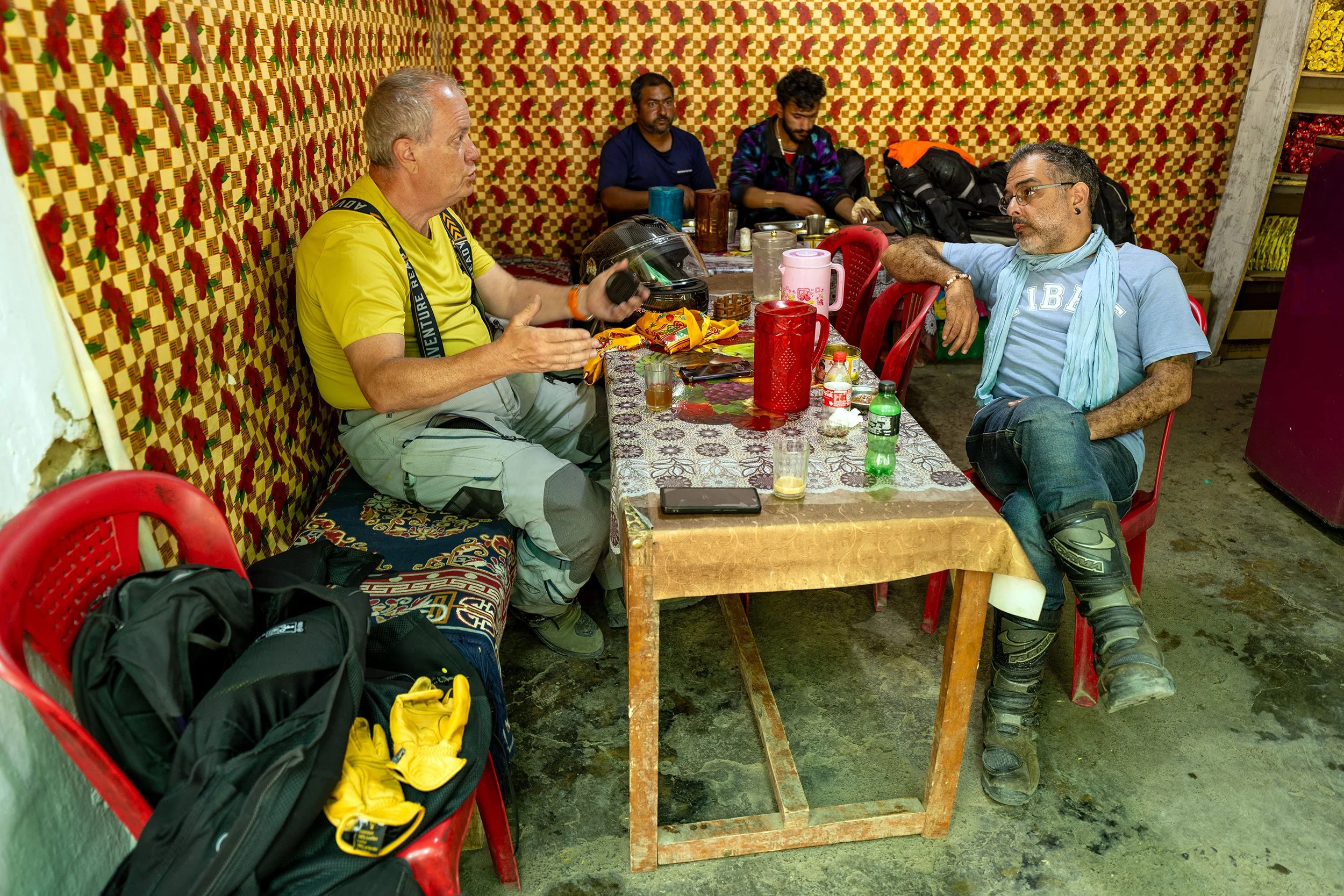 Four men sitting around a table inside a room with patterned wallpaper. The man on the left, wearing a yellow shirt, is holding a helmet and speaking to the man on the right, who is wearing a light blue shirt and glasses. The table has various drinks