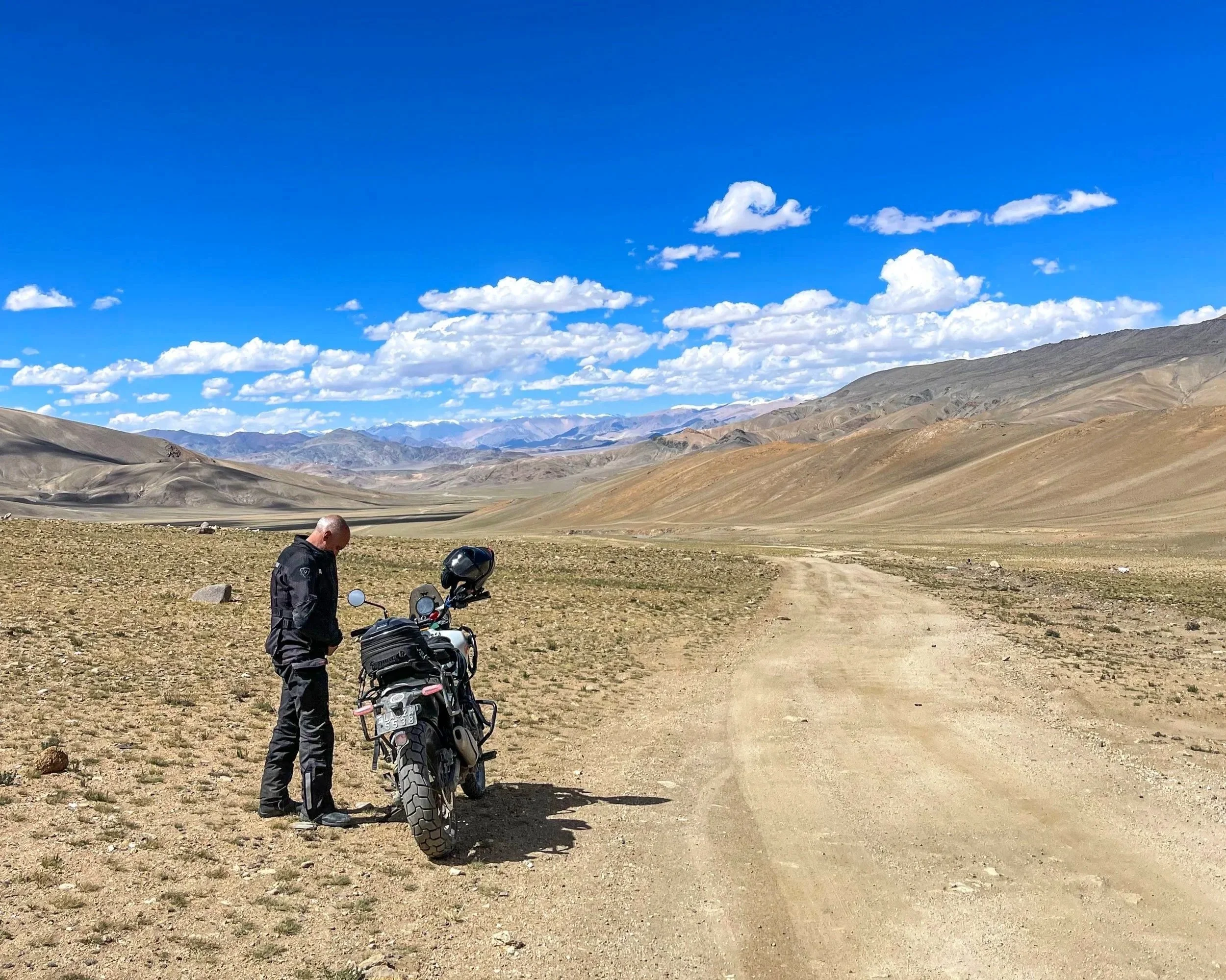 A man standing next to a motorcycle on a dirt road in a vast, arid landscape with mountains in the background under a blue sky with scattered clouds.
