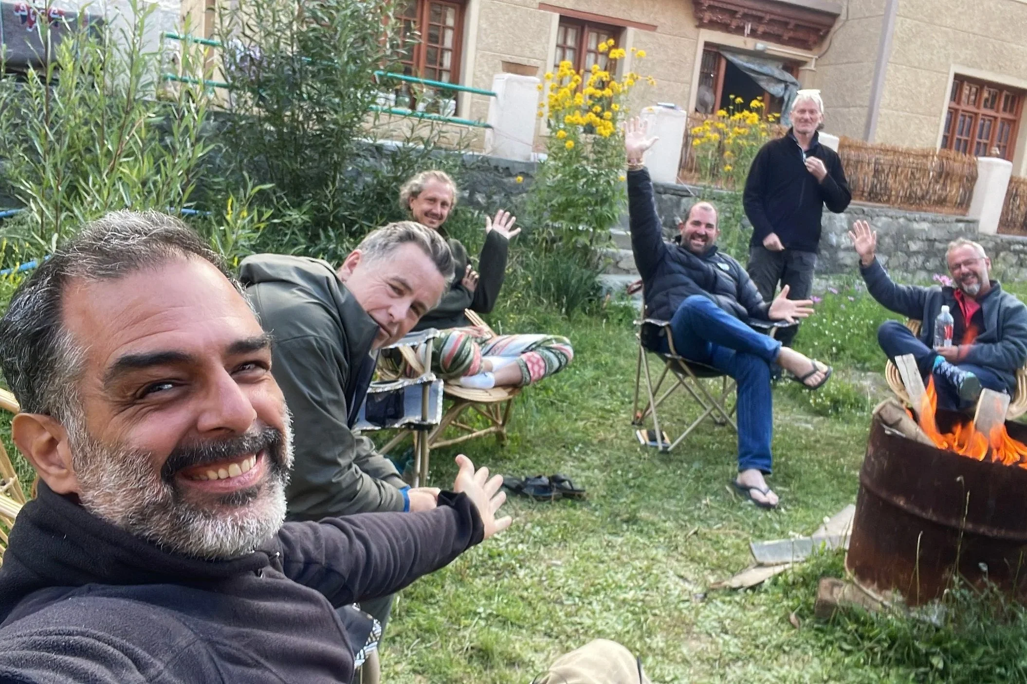 A group of several men sitting outdoors on chairs, smiling, and waving at the camera, with a fire burning in the foreground. They are in a garden setting with plants, flowers, and a building in the background.