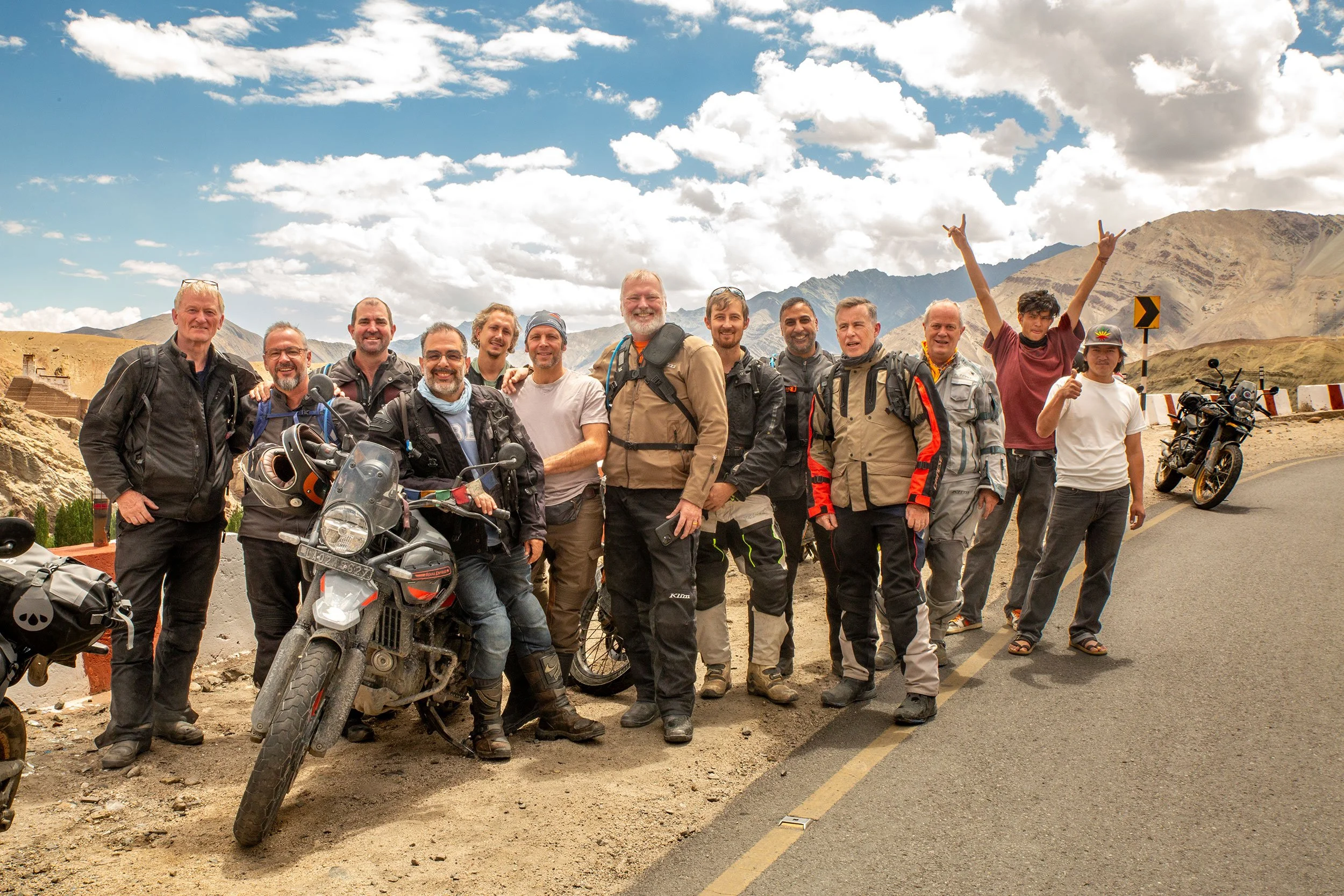 A group of twelve me, dressed in motorcycle gear, standing together outdoors in a mountainous desert landscape with clouds in the sky. There are two motorcycles and a backpack visible in the scene, and some individuals are smiling or making gestures.