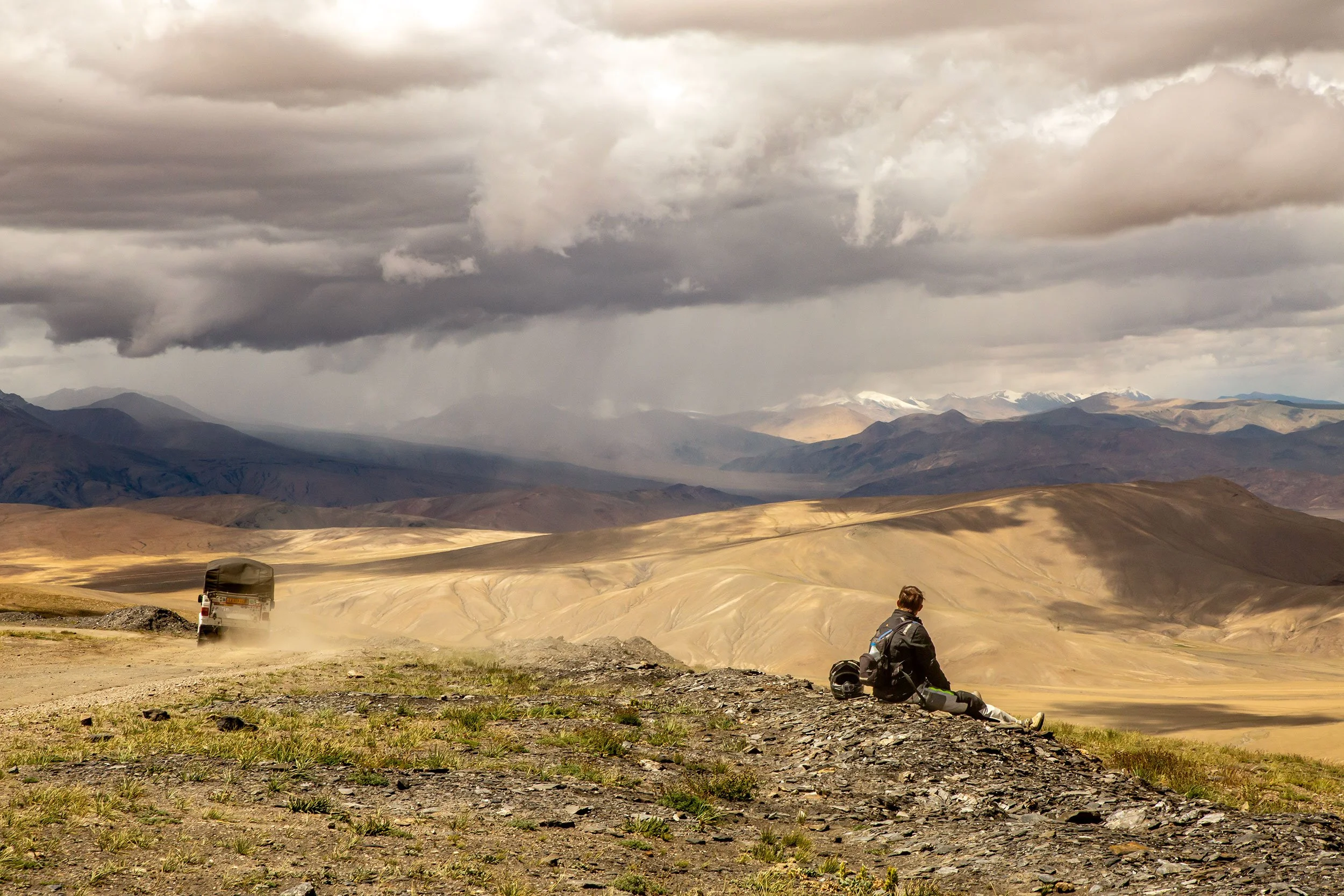 Car moves on a dirt road in a dusty mountainous landscape with cloudy sky, a man sitting on the ground, contemplating the vast lanndscape.