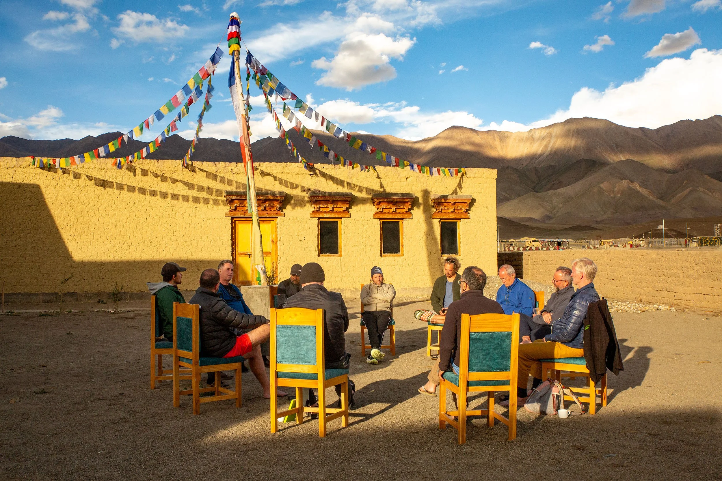 A group of men sitting outdoors in a men's circle with mountains and a blue sky in the background. Colorful prayer flags are hanging above them, attached to a tall wooden pole. They appear to be engaged in a discussion or meeting.