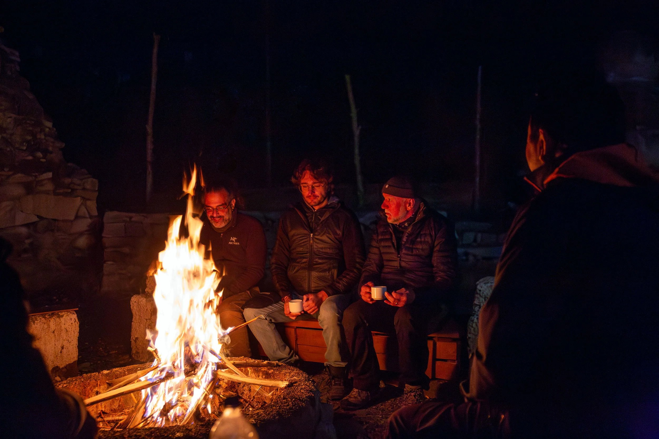 Group of people sitting around a campfire at night, holding cups and conversing.