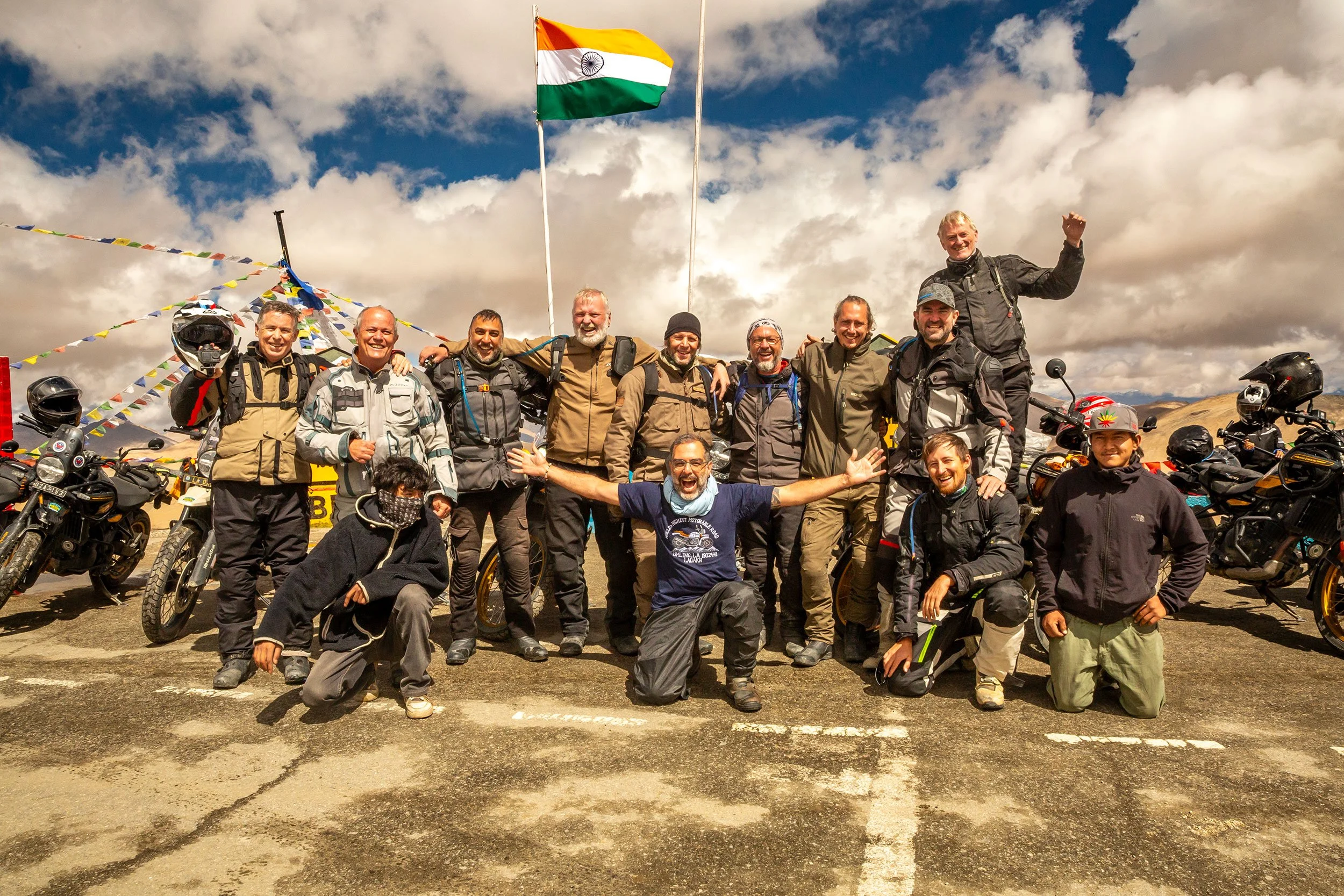 A group of happy men, dressed in riding gear, gathered together outdoors on a cloudy day, with motorcycles parked around them and an Indian flag flying in the background.