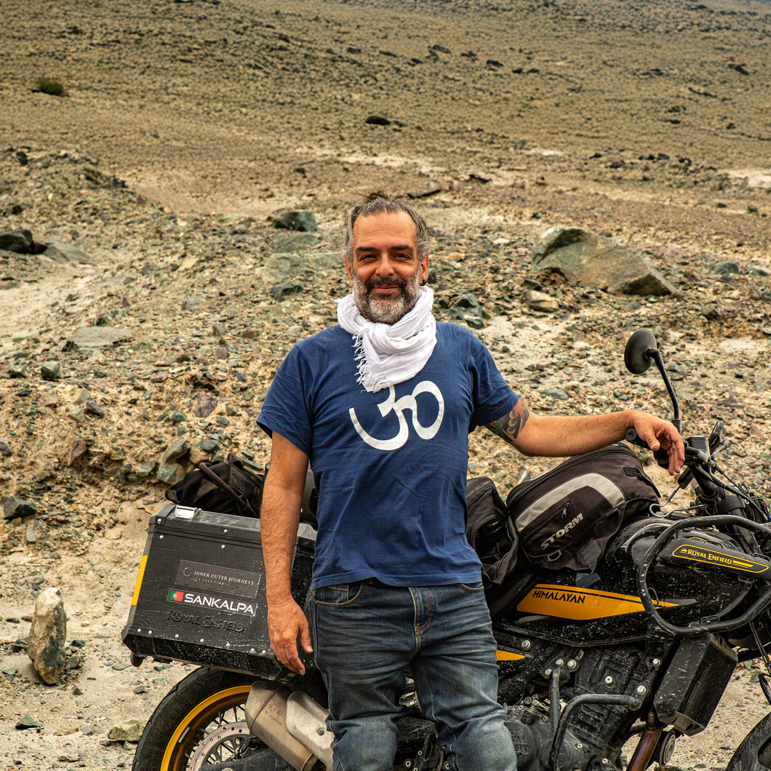 Man standing next to motorcycle in a desert landscape, wearing a blue T-shirt with an Om symbol, white scarf, and jeans.