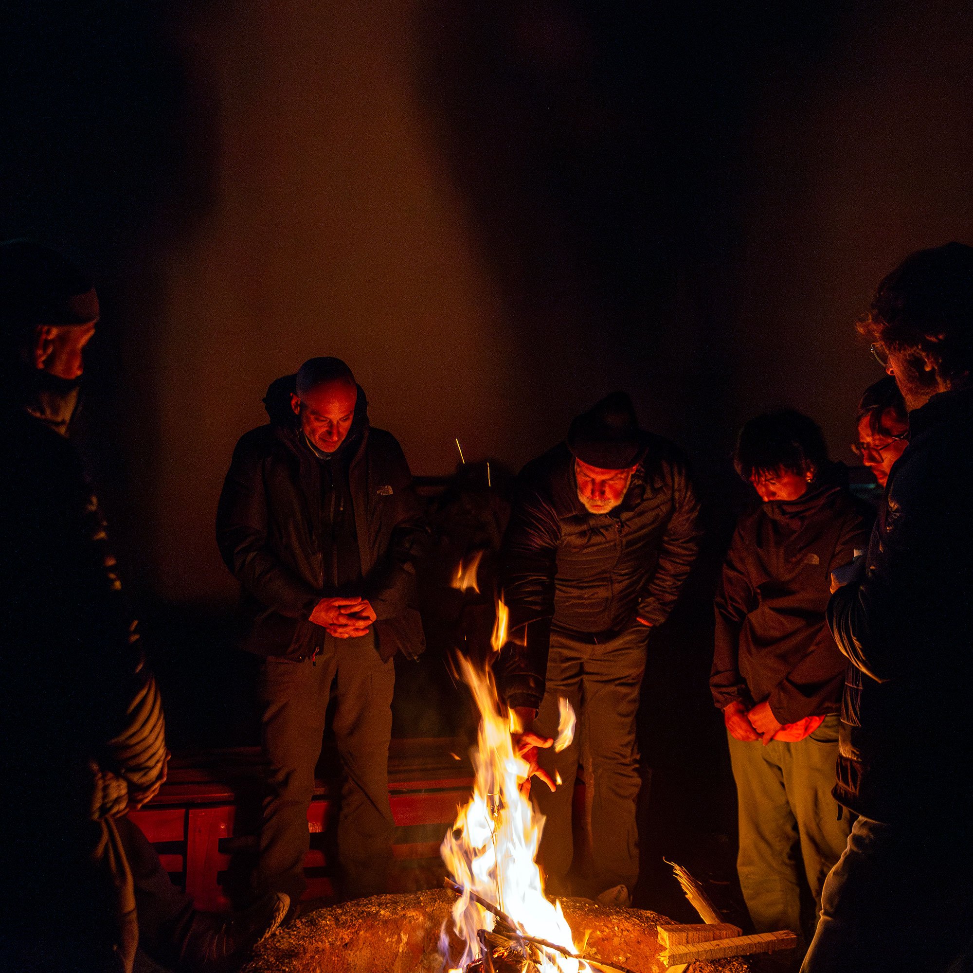 Group of men gathered for a ritual, around a campfire outdoors at night.