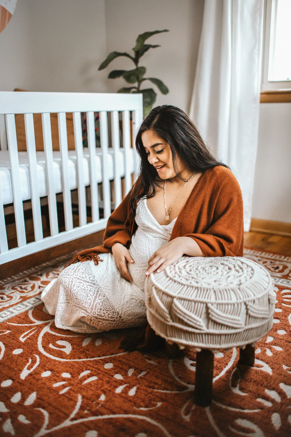 Expecting mother holding her belly leaning on a woven stool in baby's nursery, sunlight streaming in through window