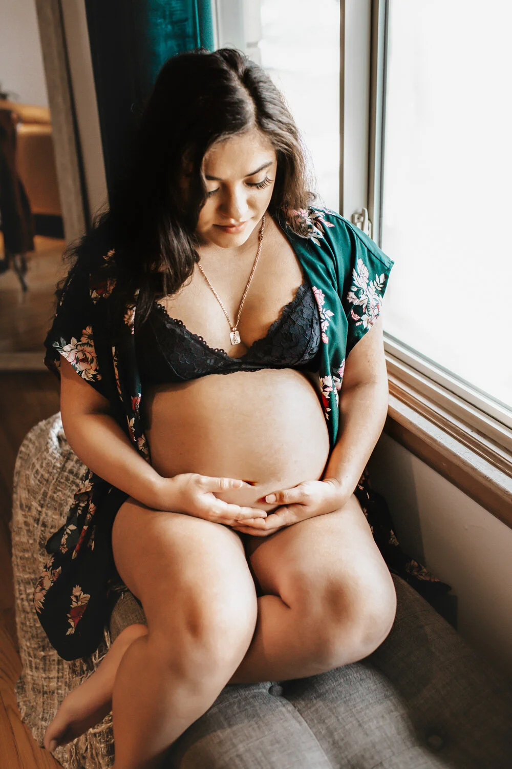 Expecting mother holding belly sitting on ottoman in living room, sunlight streaming in through window