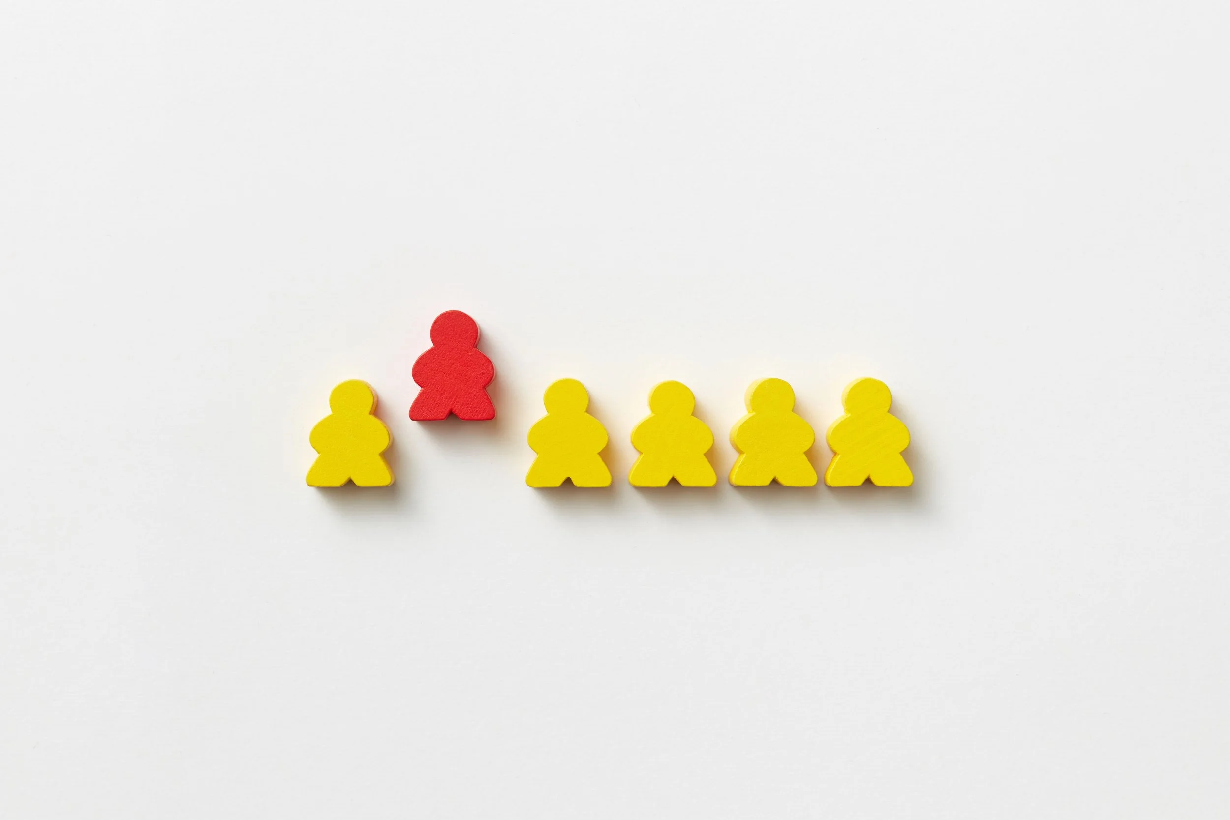 Six human shaped cut outs lined up horizontally. Five colored yellow and one colored red cut pushed up vertically out of line.