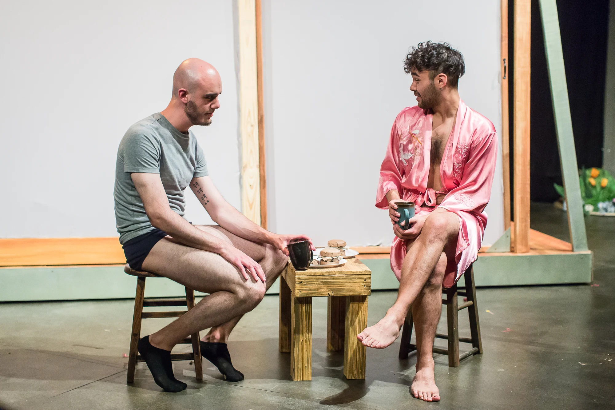An image of two men sit facing each other on chairs, sharing a small table with cups and snacks, against a bare wooden structure, engaging in conversation.