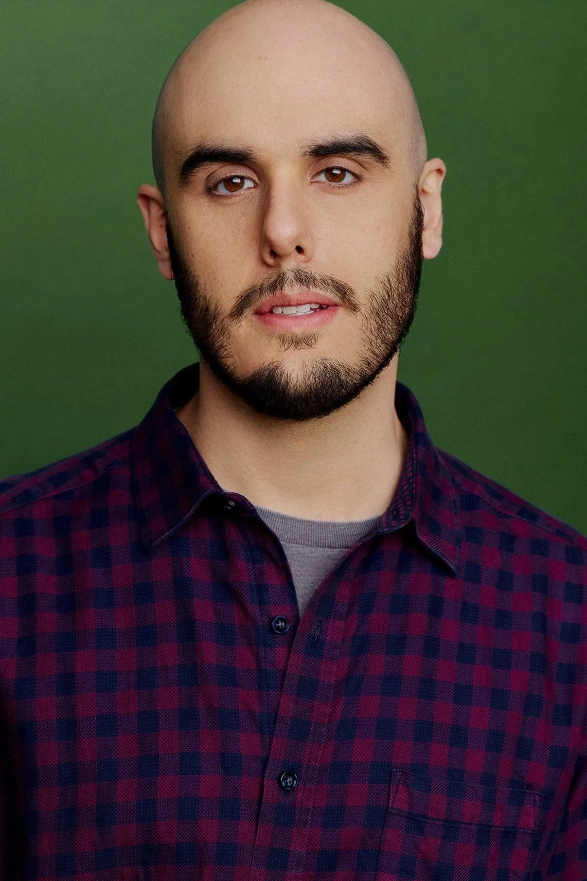 A headshot portrait of a young man with a beard and a shaved head, wearing a maroon and navy blue checkered button-up over a charcoal gray shirt, posing against a green background.
