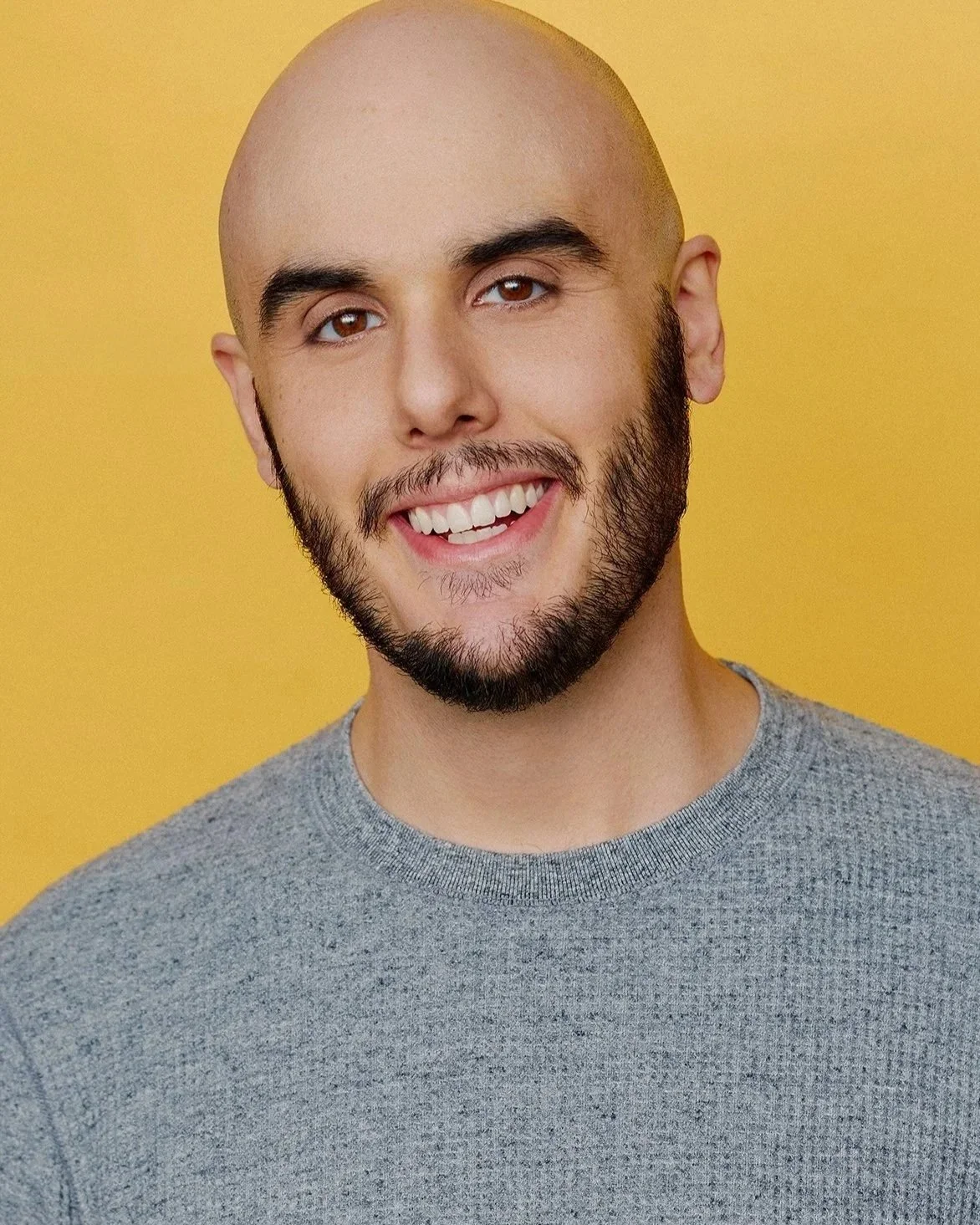 A headshot of a smiling young man with a beard and a shaved head, wearing a gray waffle-print shirt against a yellow-orange background.