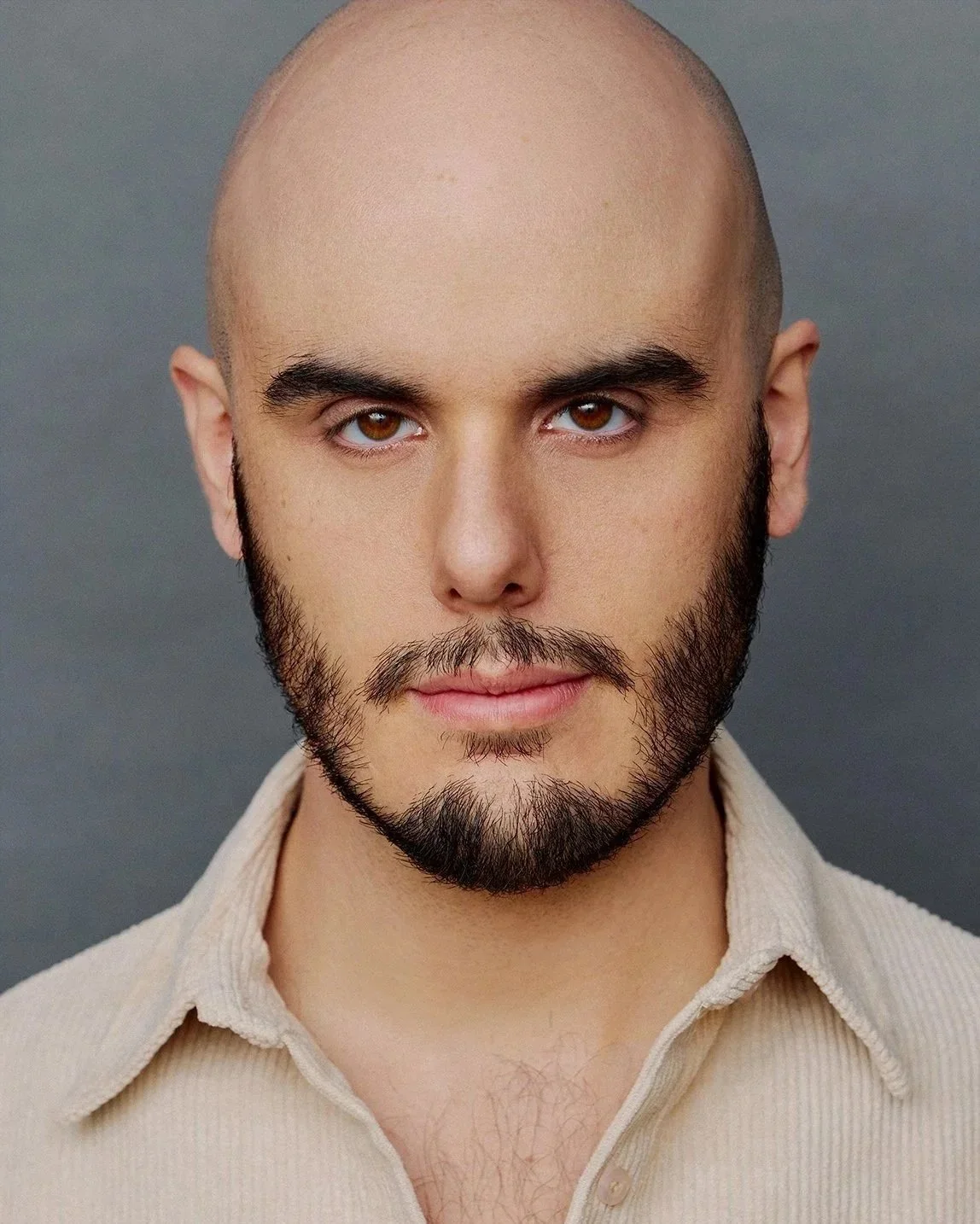 A headshot portrait of a man with a shaved head, brown eyes, and a beard, wearing a beige corduroy button-up against a charcoal gray background. He has a serious look on his face.