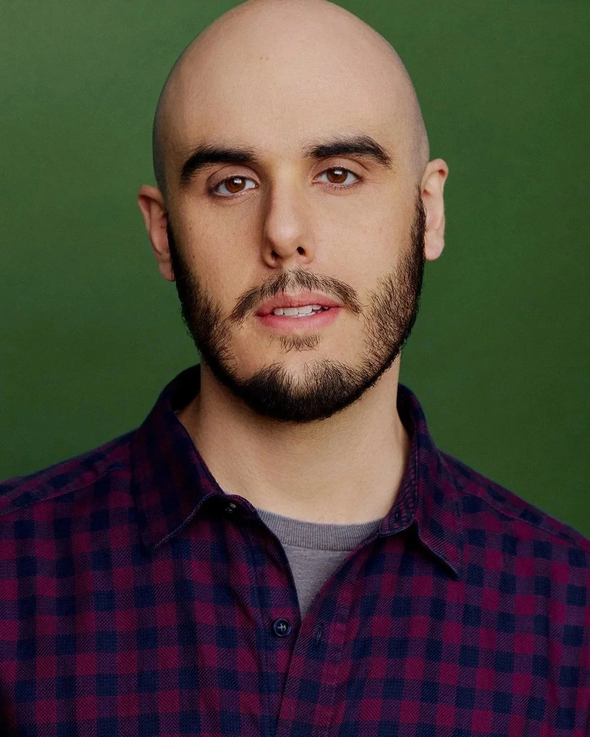 A headshot portrait of a young man with a beard and a shaved head, wearing a maroon and navy blue checkered button-up over a charcoal gray shirt, posing against a green background.