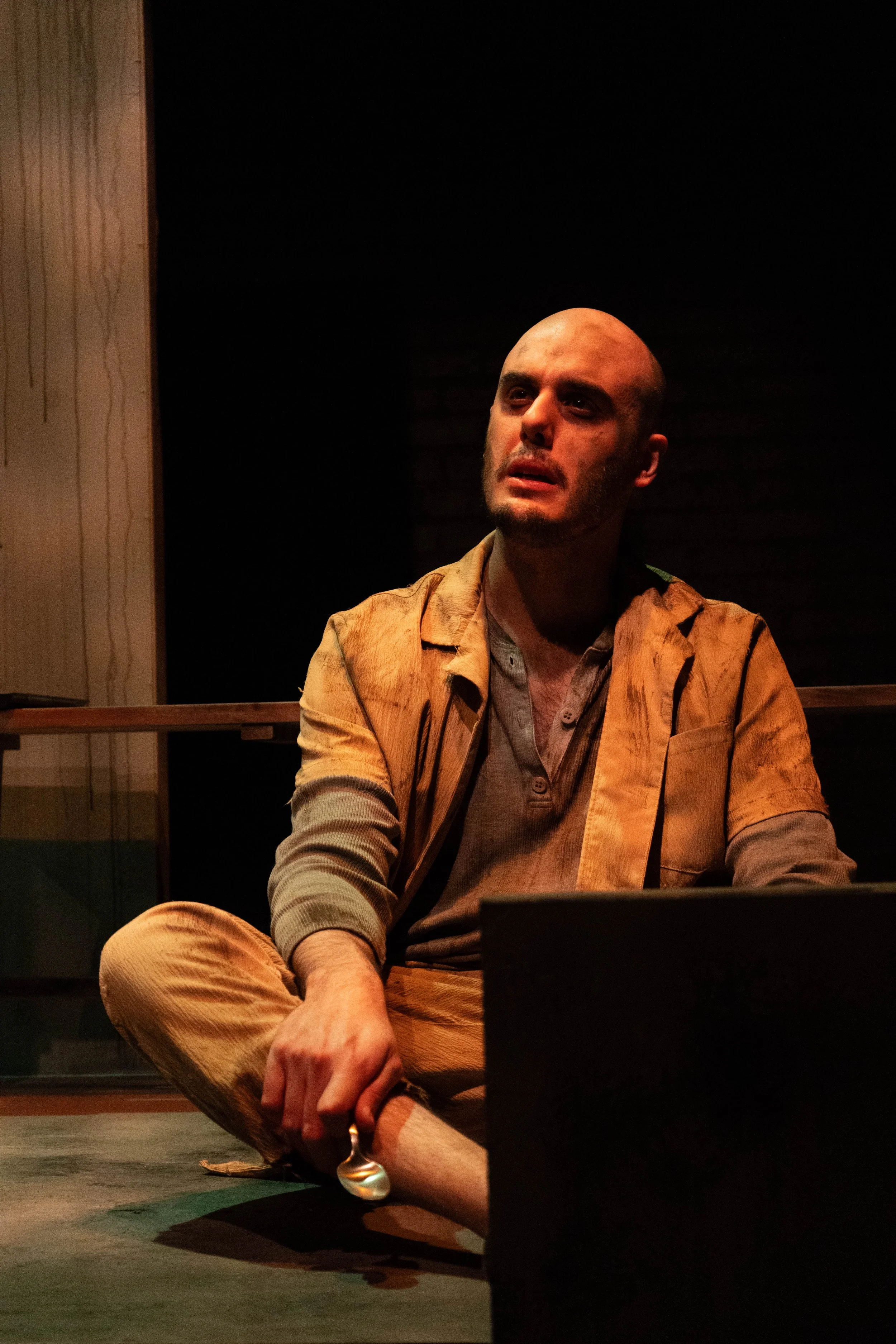 A man with a bald head and a beard sits on the floor in a dimly lit room, looking upwards with a distressed expression. He is wearing a tan jacket over a gray shirt and tan pants, holding a spoon in his left hand.