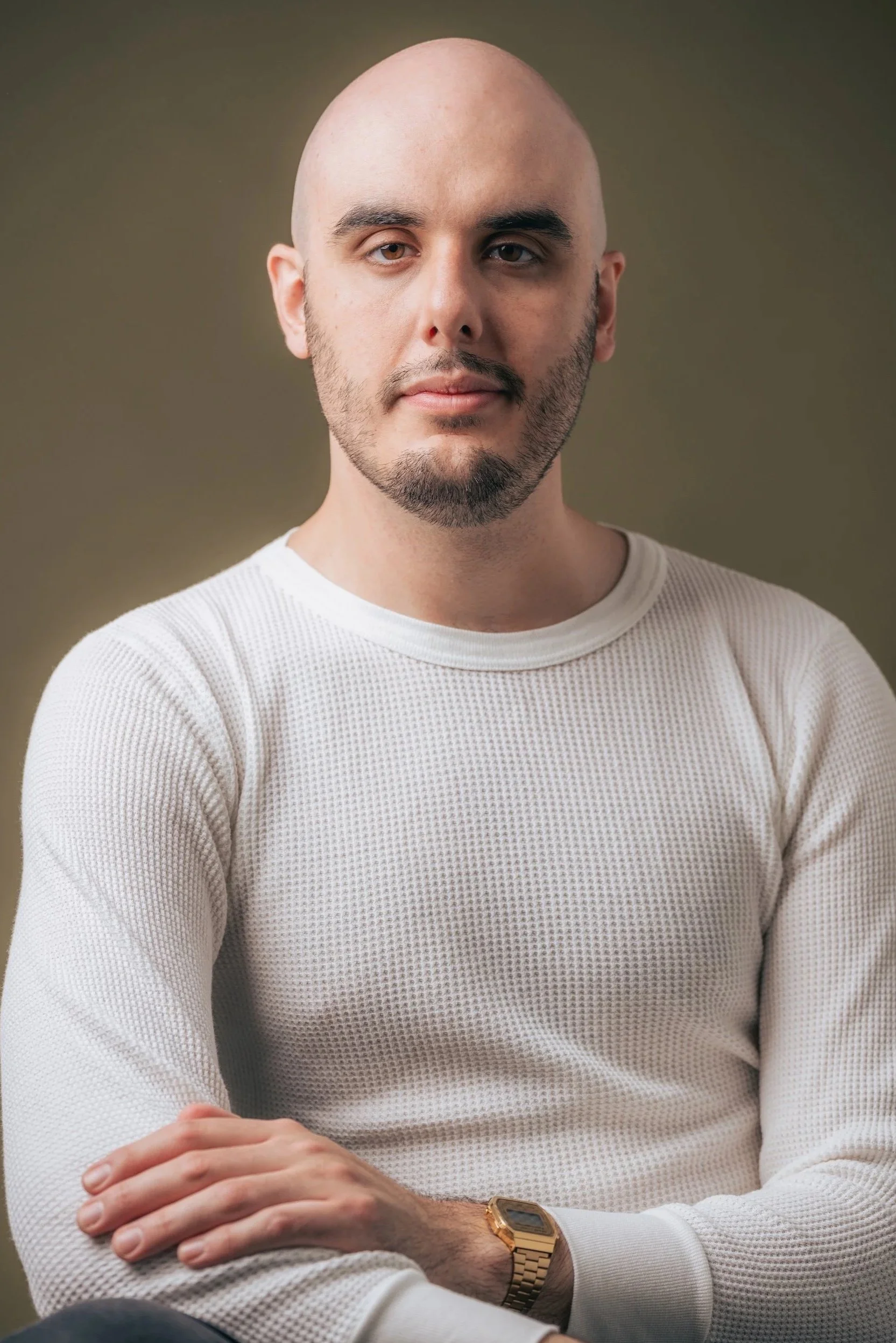 A portrait of a young man with a bald head, beard, and light skin, wearing a white textured long-sleeve shirt and a gold wristwatch, sitting against a brown plain background.