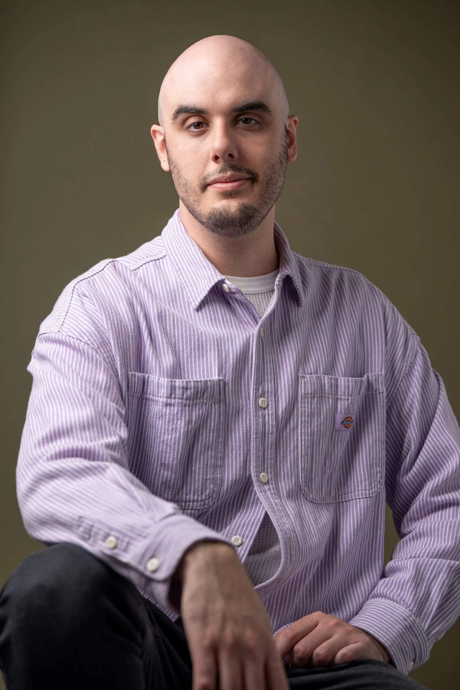 A young man with a bald head and beard, wearing a light lilac striped button-down shirt sitting with his arm resting on his knee against a plain, muted background.